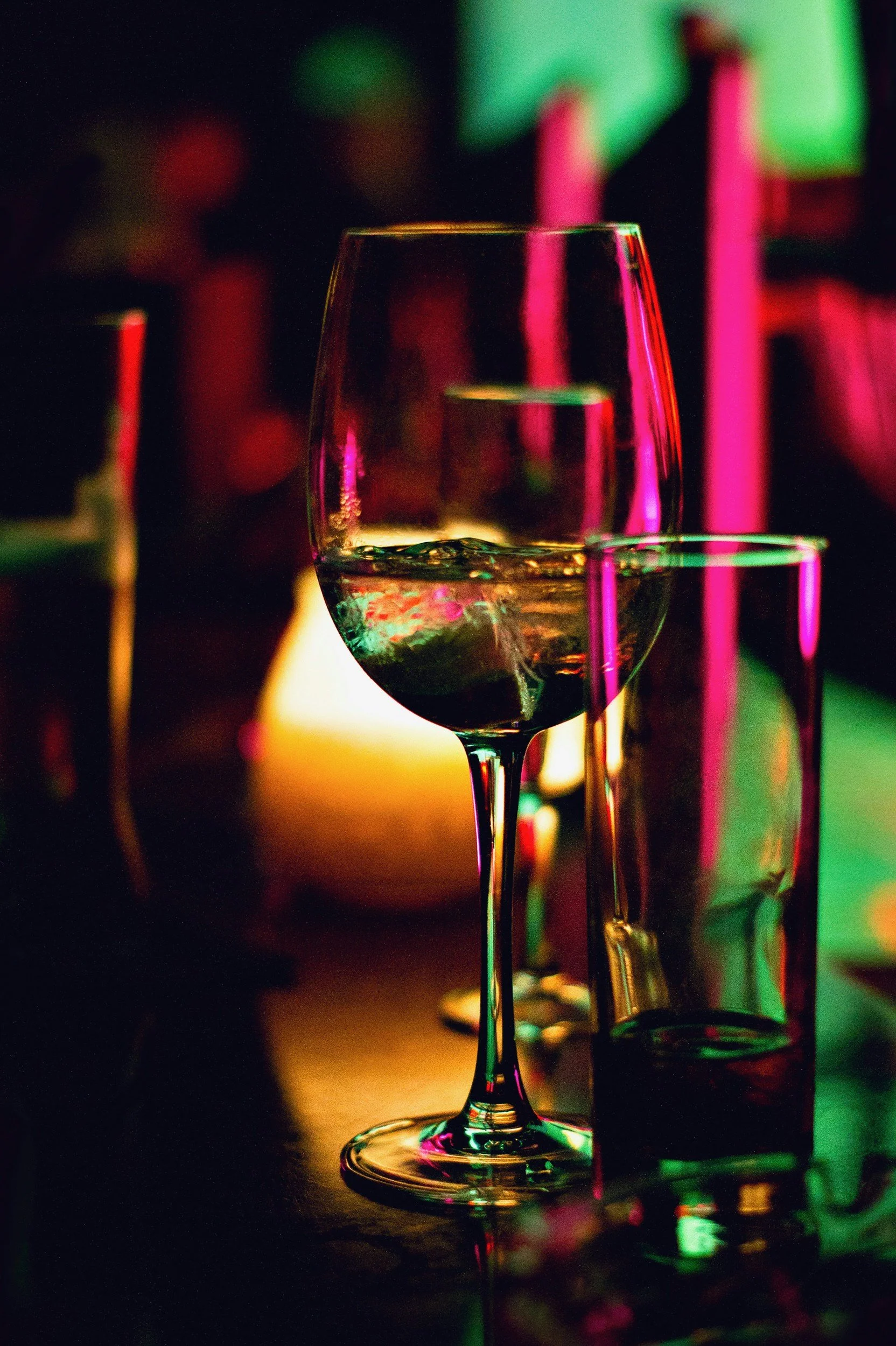 A glass of wine on a dark surface with colorful neon lights reflecting on it, creating a vibrant and moody atmosphere.