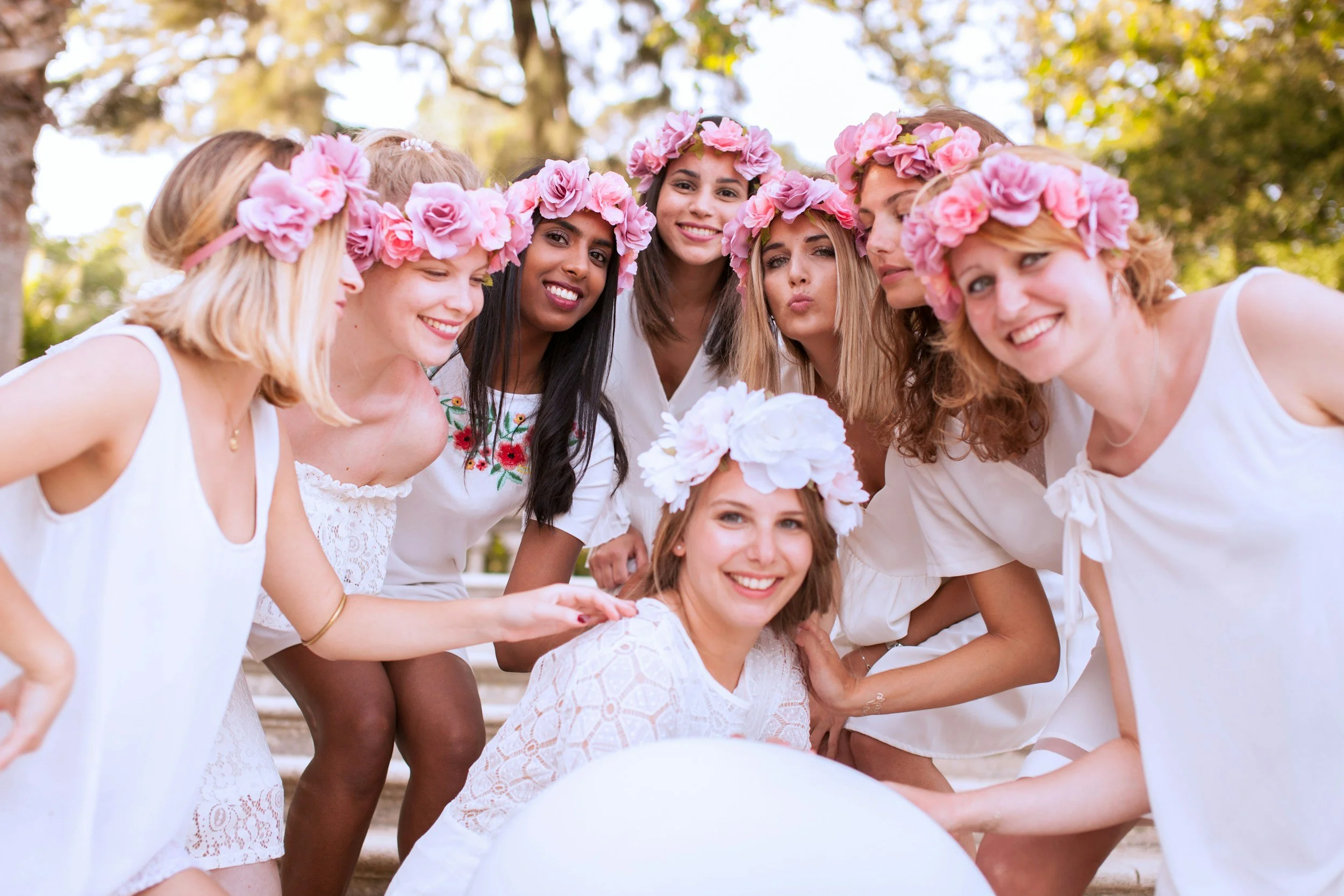 Young women wearing white dresses and pink floral crowns, gathered outdoors, smiling and taking a group photo.
