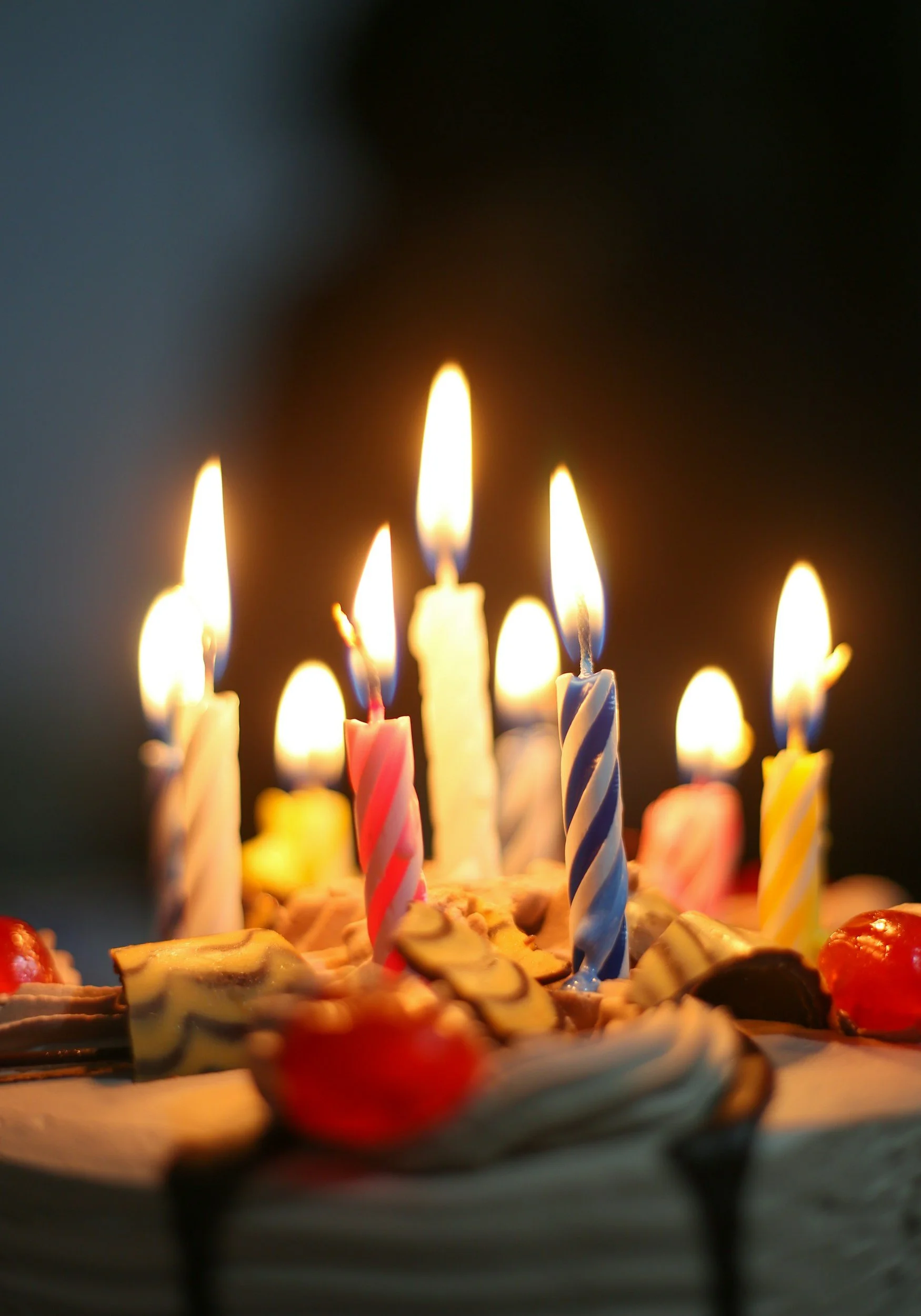 Close-up of a birthday cake with multiple lit candles of various colors, with a dark background.