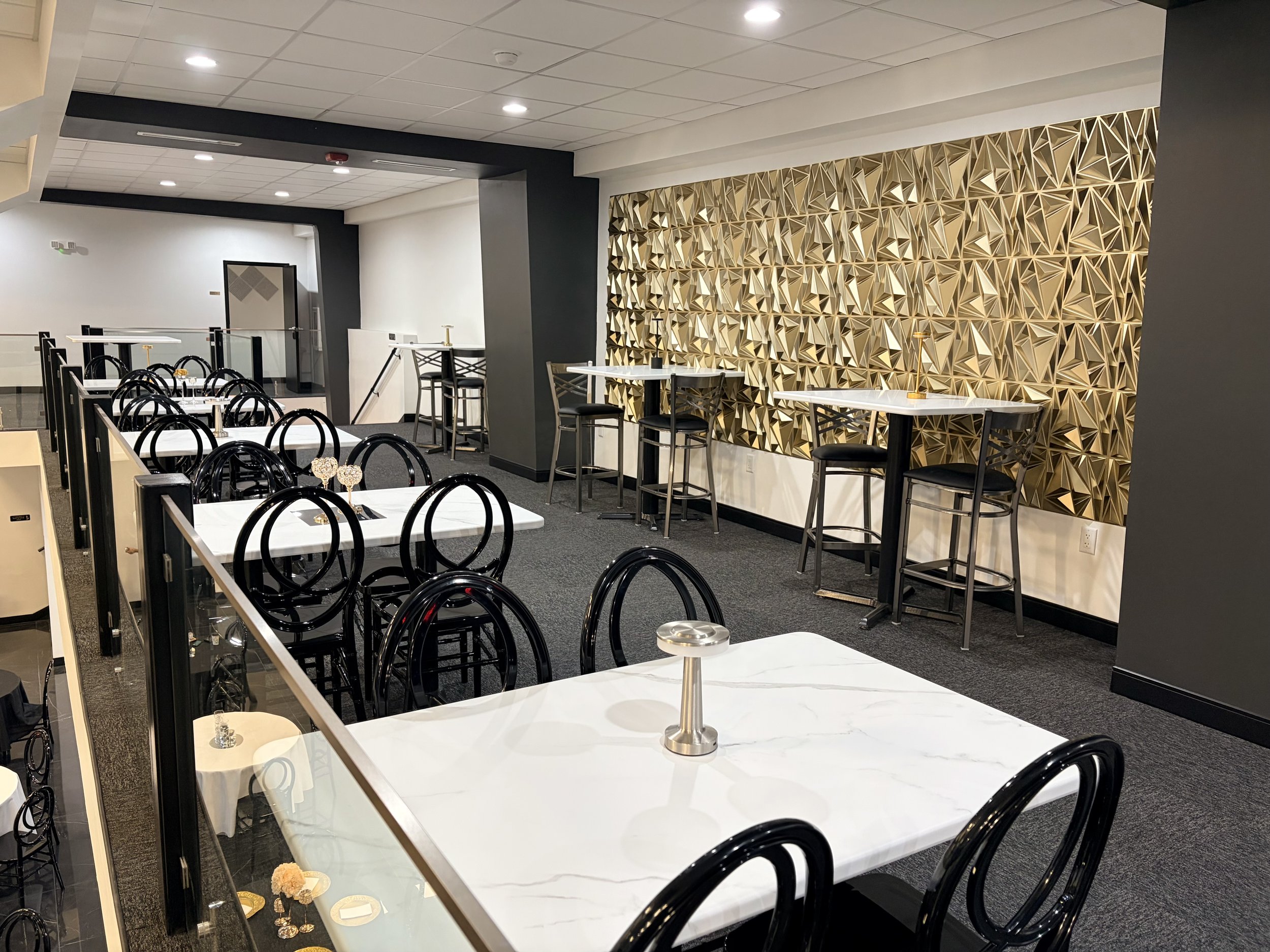 A modern indoor dining area featuring black chairs, white marble tables with decorative items, and a striking gold geometric wall accent. There are also bar-height tables with black chairs and a staircase in the background.