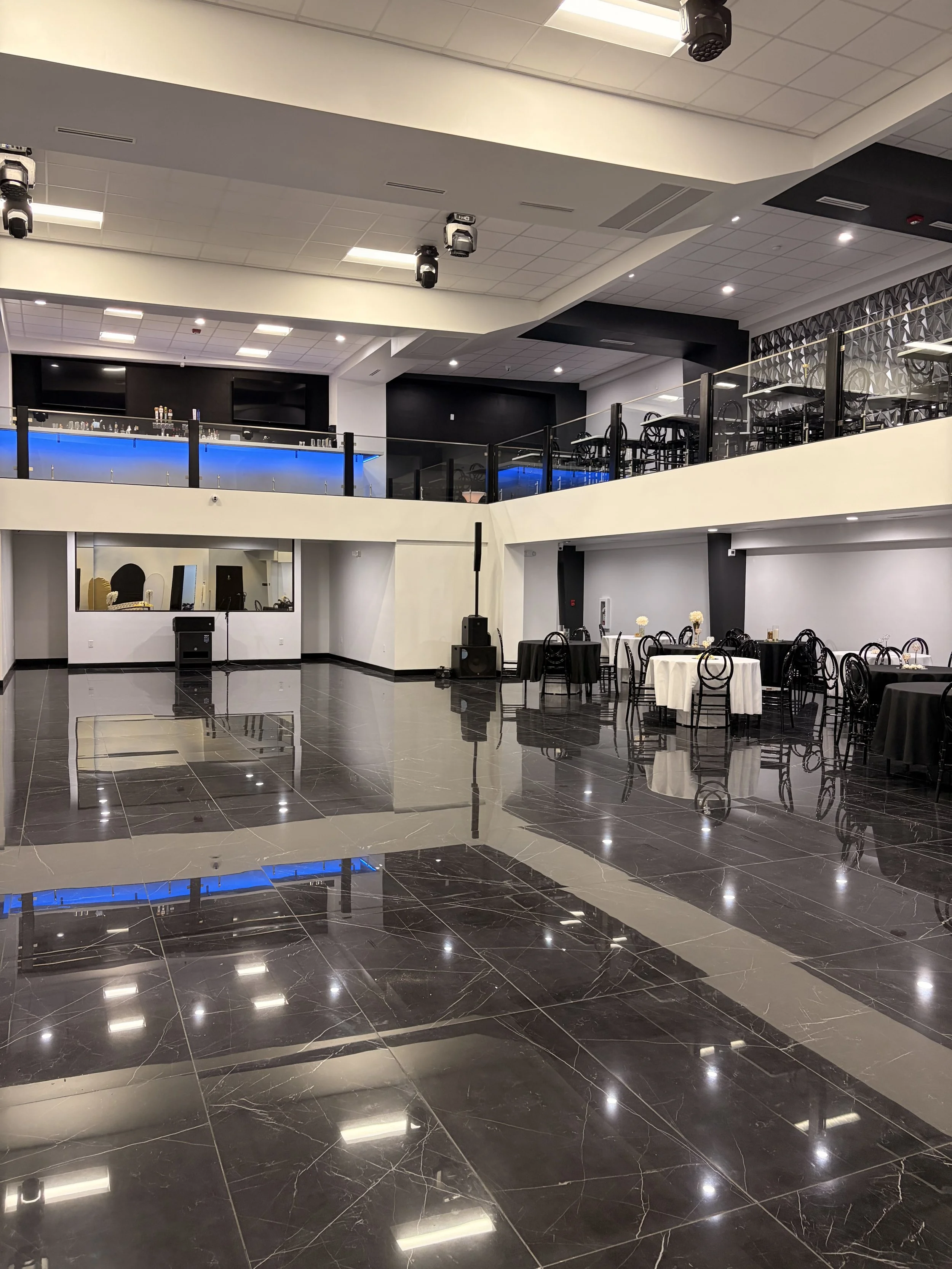 An empty event space or banquet hall with black and white decor, tables with white tablecloths and black chairs, a second-floor balcony with tables and chairs, and a black and blue illuminated bar area.