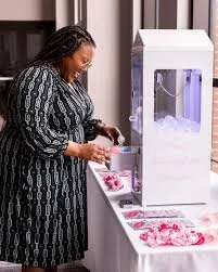 A woman standing at a table with a 3D printing machine, preparing to print with pink filament. The table has pink objects or materials.