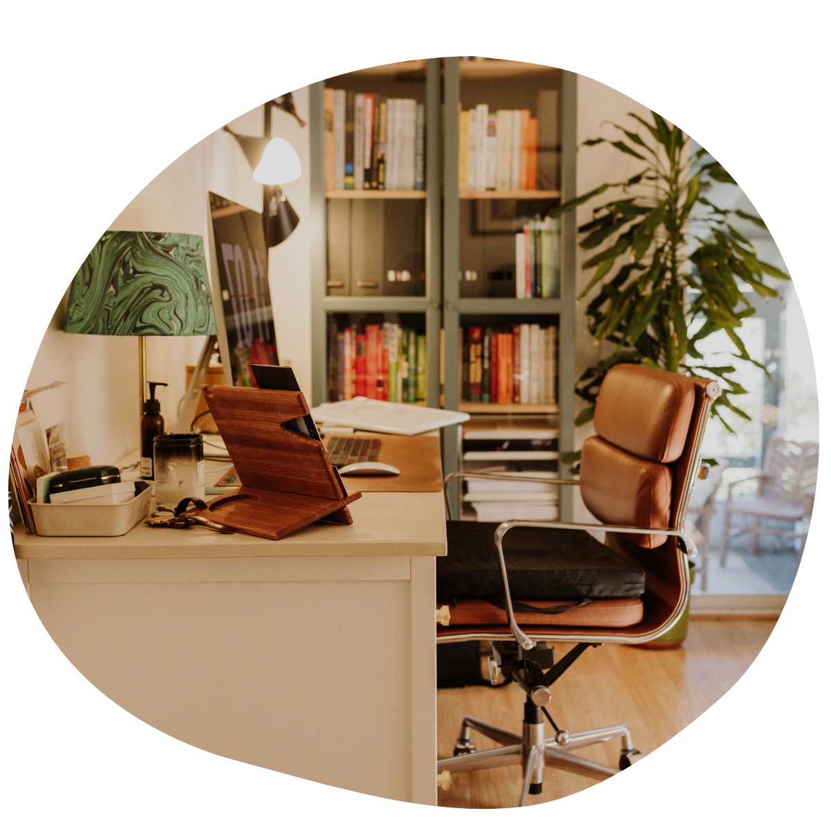 Home office with desk, brown leather chair, bookshelf, green plant, lamp, and various office supplies.