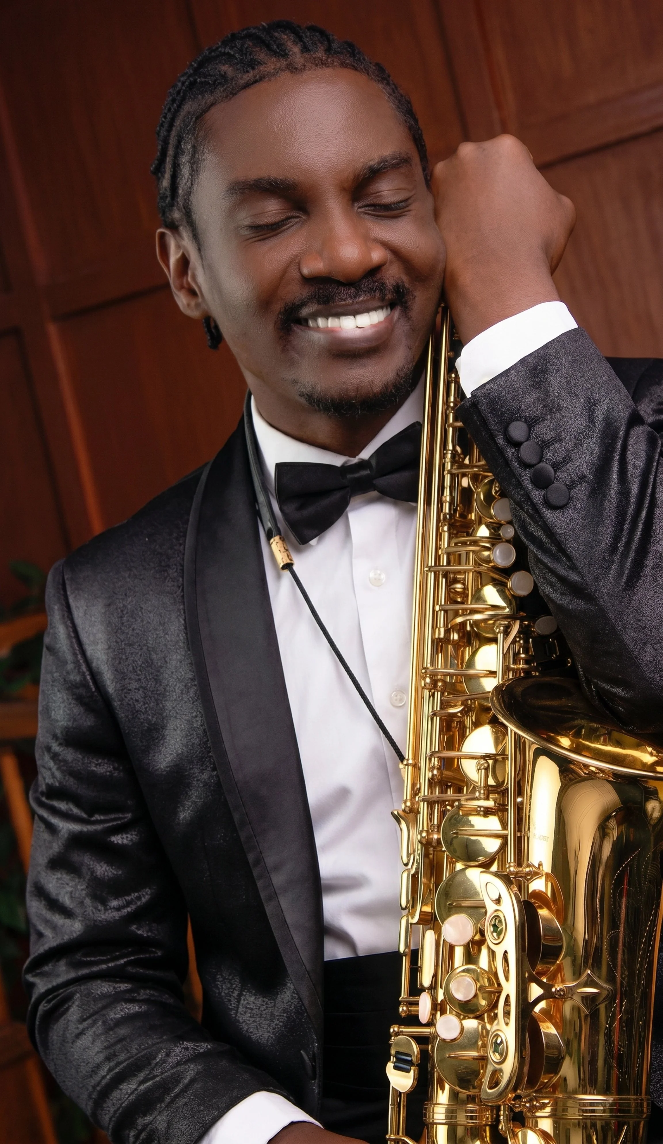 A man with dreadlocks wearing a tuxedo, smiling, holding a gold saxophone, standing indoors against a wooden background.