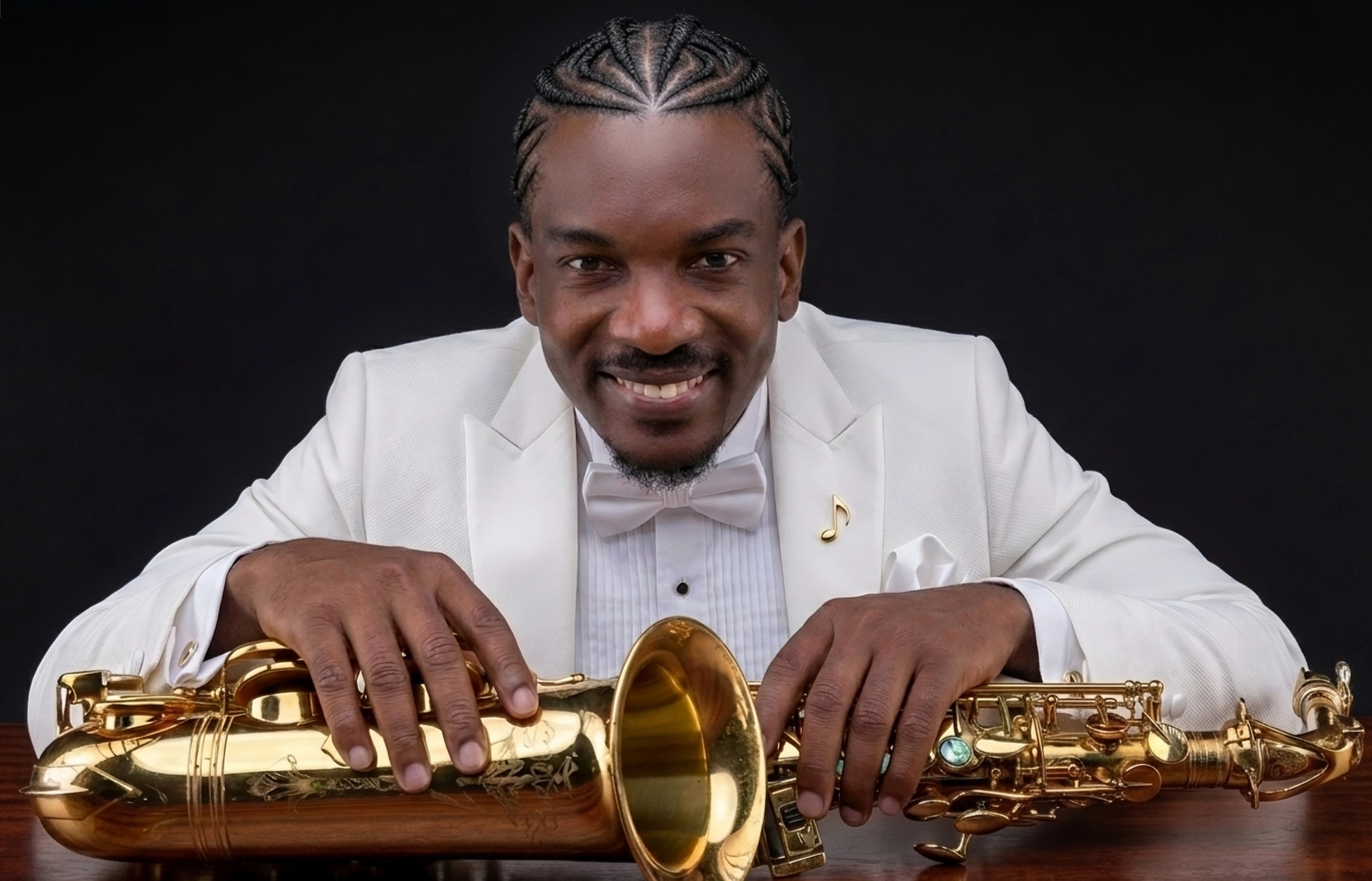 A man in a white tuxedo with a bow tie and musical note pin is holding a gold saxophone and smiling at the camera against a black background.