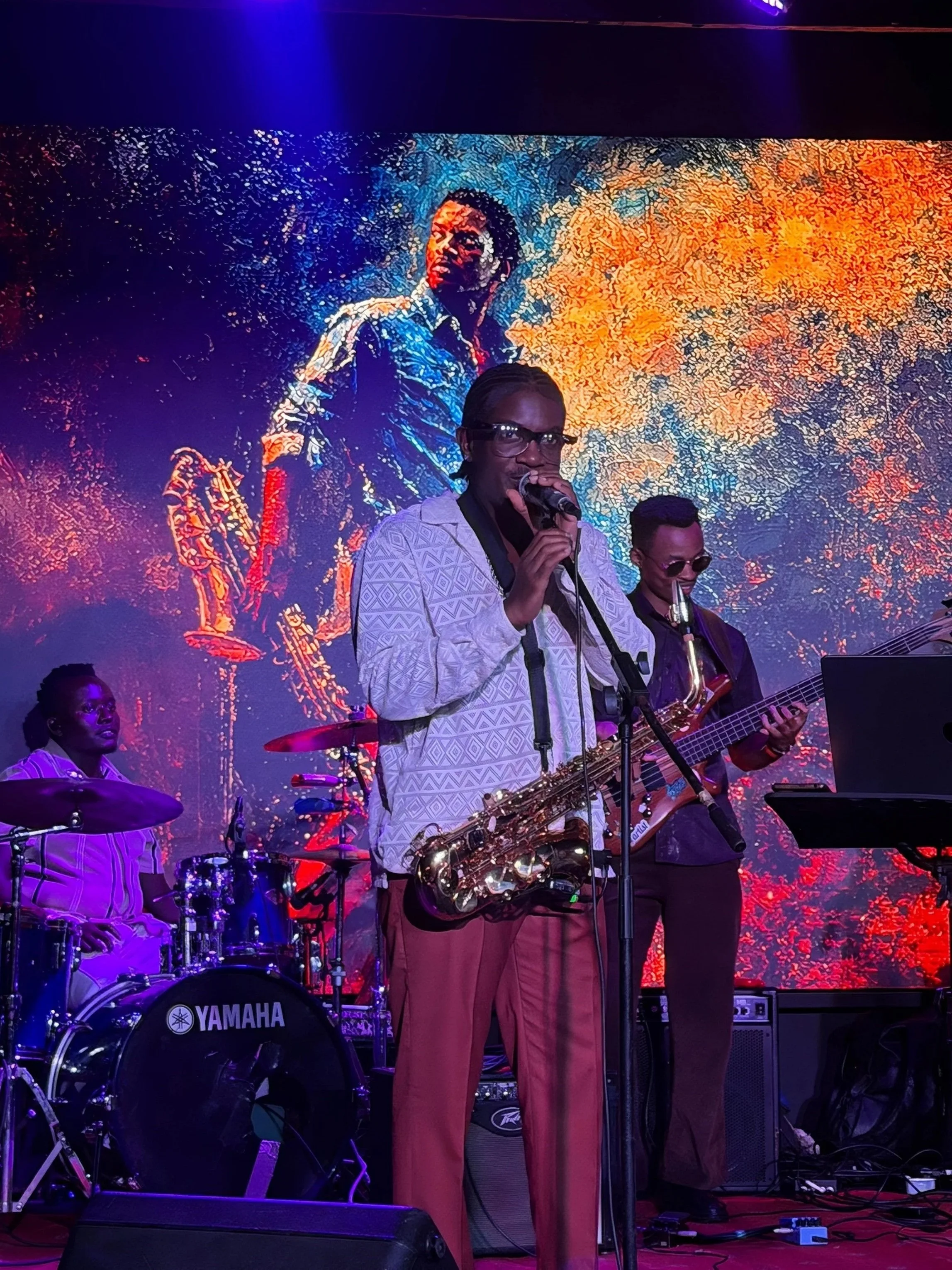 A band performing live on stage with a woman singing into a microphone, a man playing a saxophone, a drummer, and a bassist, with a colorful digital background.