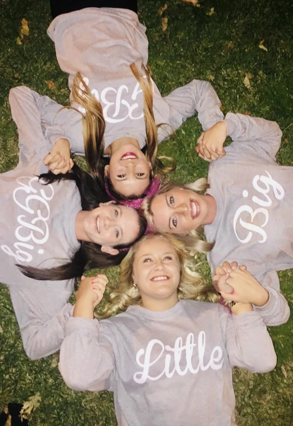 Five women are lying on their backs on grass in a circle, holding hands, with heads close together. They are smiling and looking up at the camera. They are all wearing matching gray shirts with white cursive text; one shirt says "Little" and the others say "Boss." The women have different hair colors and styles, and the background is grass with some small yellow leaves.