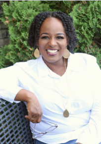 Woman with curly hair in a white blouse sitting outdoors in front of greenery, smiling.