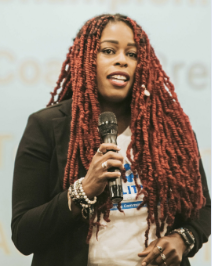 Woman with long red braided hair speaking into a microphone at an event.