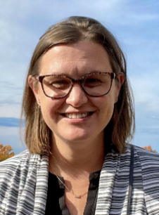 Woman with shoulder-length brown hair, wearing glasses and a black and white striped jacket, smiling outdoors with a blue sky and body of water in the background.