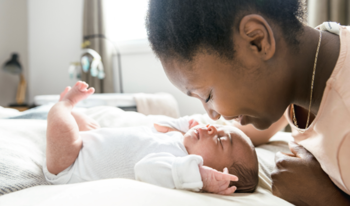 A woman with short curly hair leaning over a newborn baby lying on a bed, both smiling at each other.
