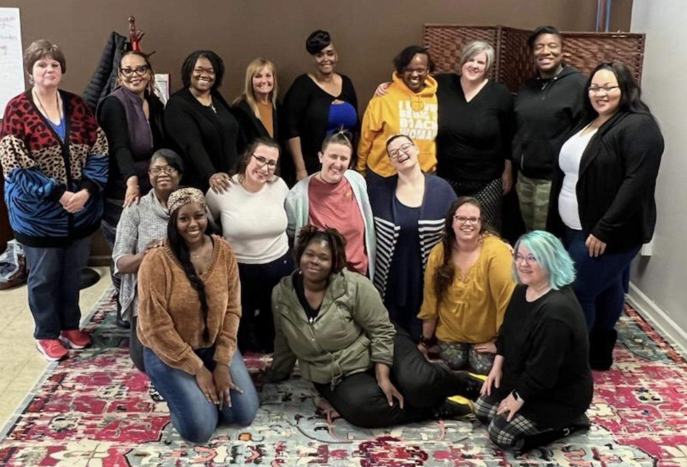 Group of women of diverse ethnicities standing and sitting on a colorful rug, smiling for a photo indoors.