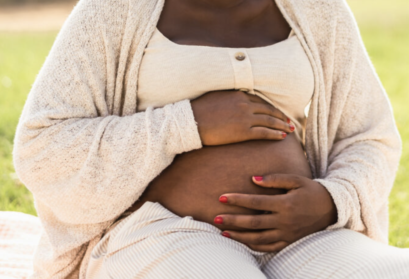 A pregnant woman sitting outdoors with hands on her belly, wearing a beige cardigan and a cream-colored top.