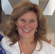 A smiling woman with shoulder-length wavy reddish-brown hair, wearing a white top and earrings, indoors with a ceiling and some furniture visible in the background.