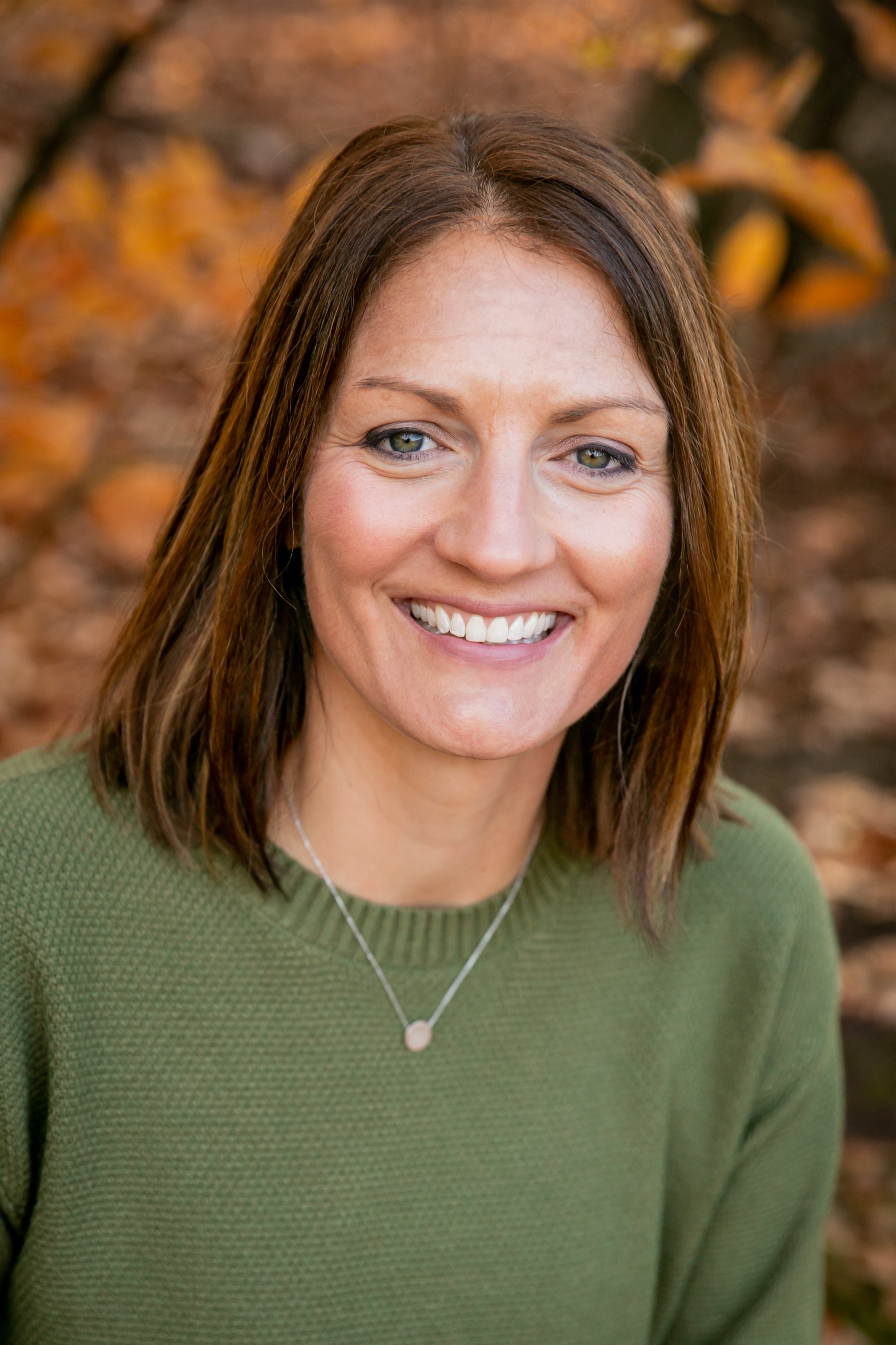 A woman smiling outdoors with autumn leaves in the background, wearing a green sweater and a silver necklace.