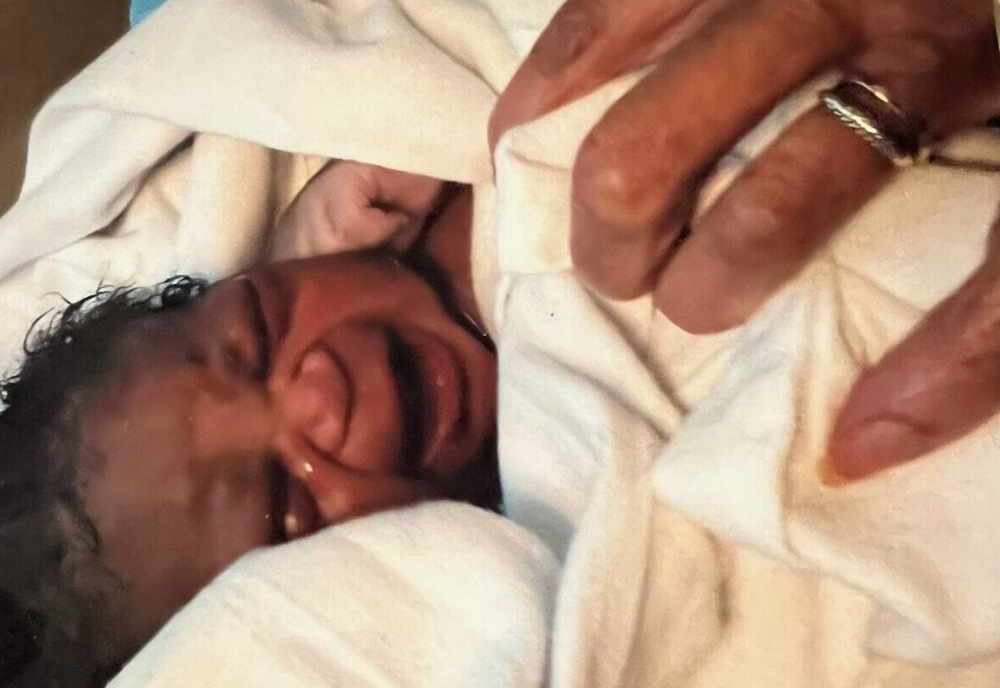 A person lying down, smiling, with their hand near their face, wearing rings, on a white fabric surface.