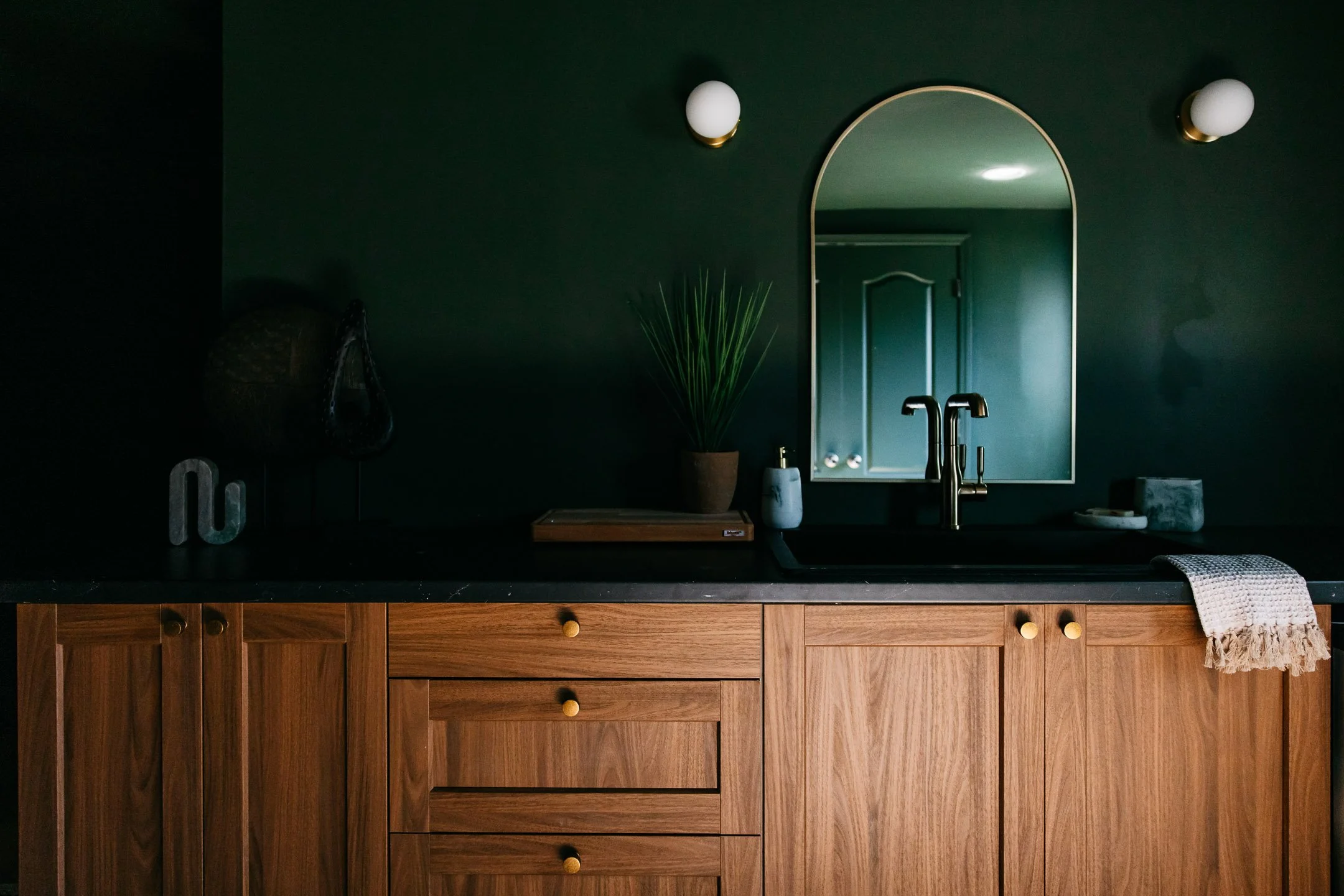 Kitchen with dark green wall, mirror, two wall sconces, wooden cabinets, and kitchen items on black countertop.