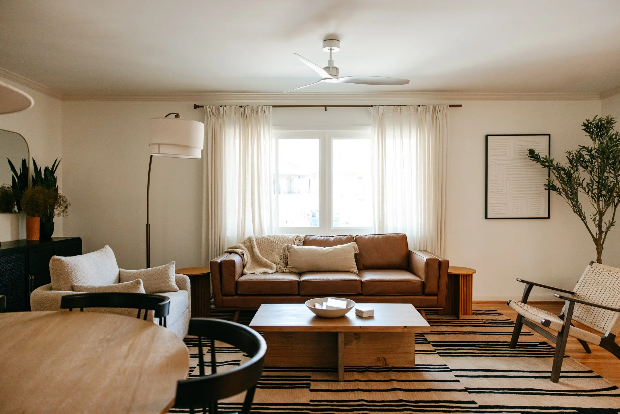 Living room with beige and leather furniture, a wooden coffee table, potted plants, and a window with cream curtains.