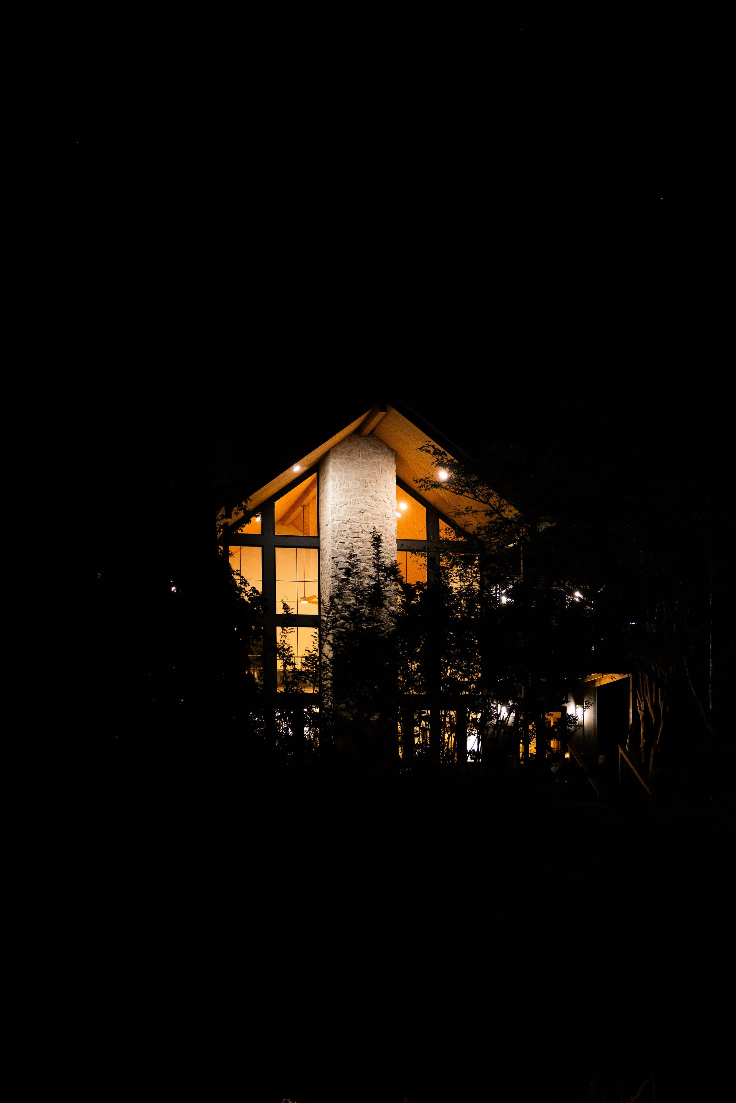 Night view of a modern house with large glass windows and a stone chimney, illuminated from inside, surrounded by trees.