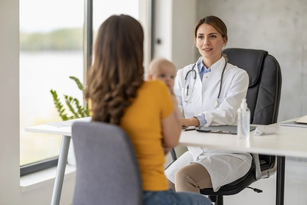 A young female doctor with a stethoscope talking to a mother holding a baby in a medical office.