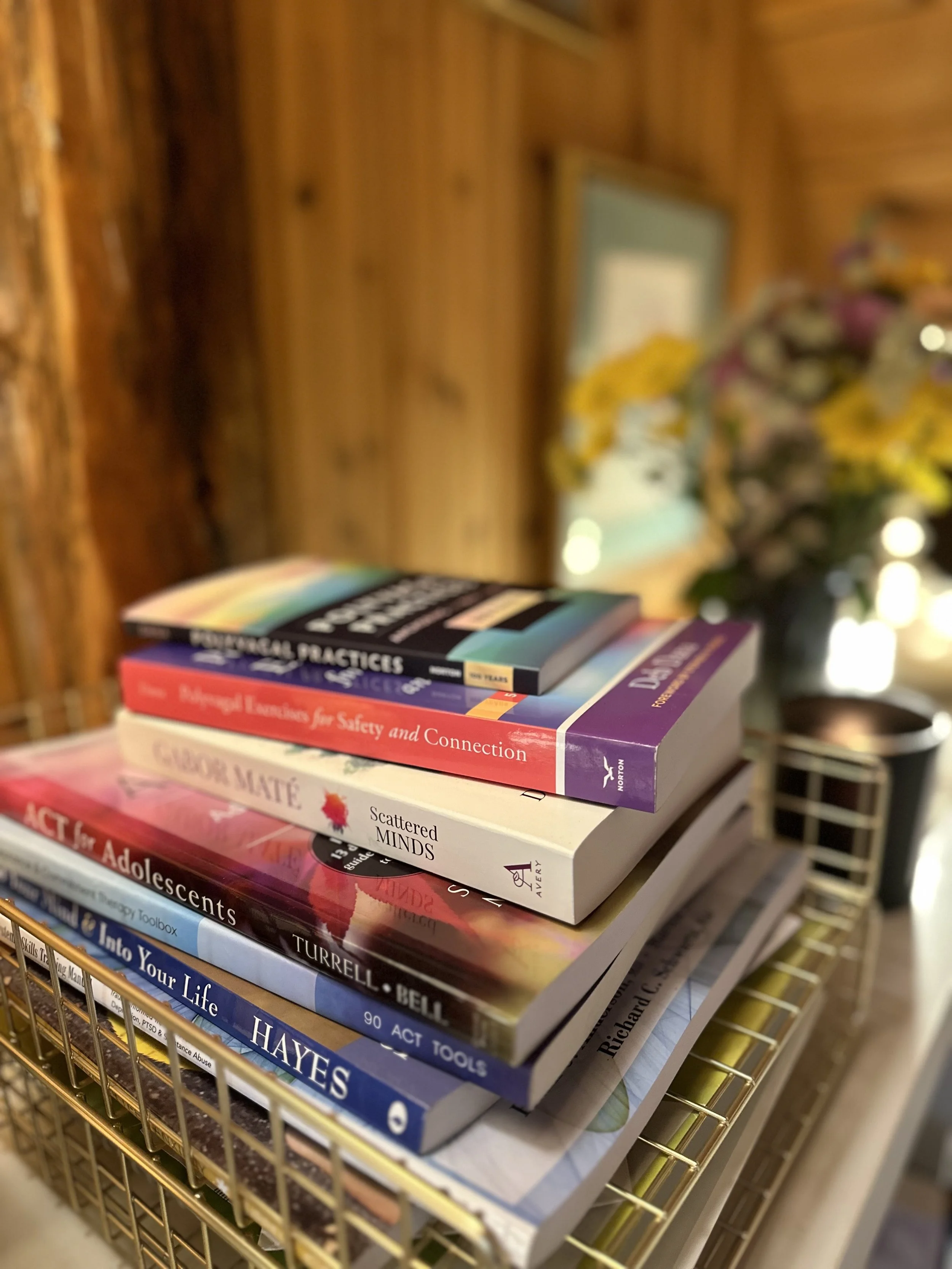 Stack of books with titles related to psychology, adolescence, and mental health on a gold-colored wire shelf, with a wooden wall and a bouquet of flowers in the background.