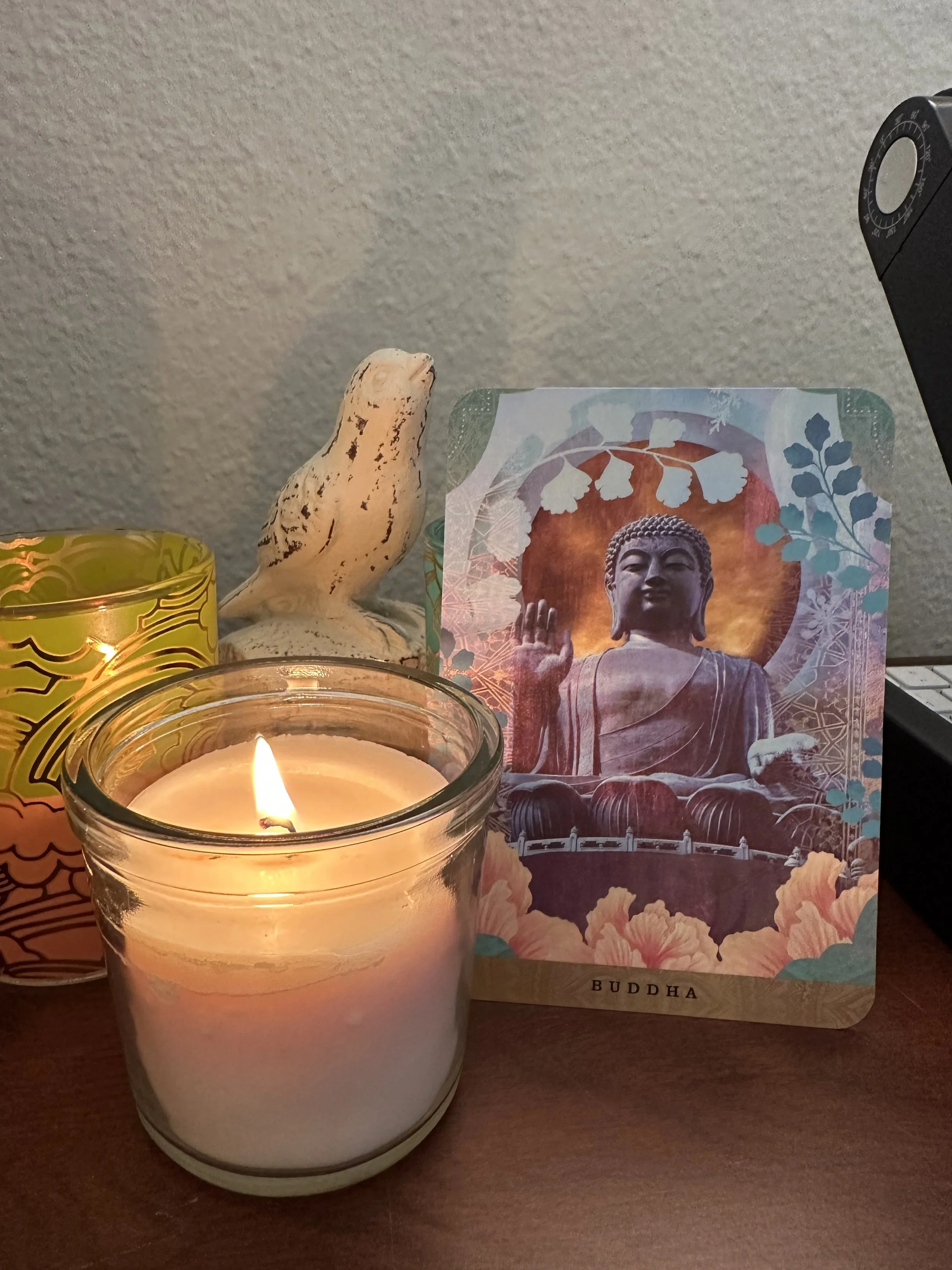 A lit candle in a glass container, a paper with a Buddha image, a small carved Buddha sculpture, and a yellow decorative candle holder are arranged on a wooden surface.