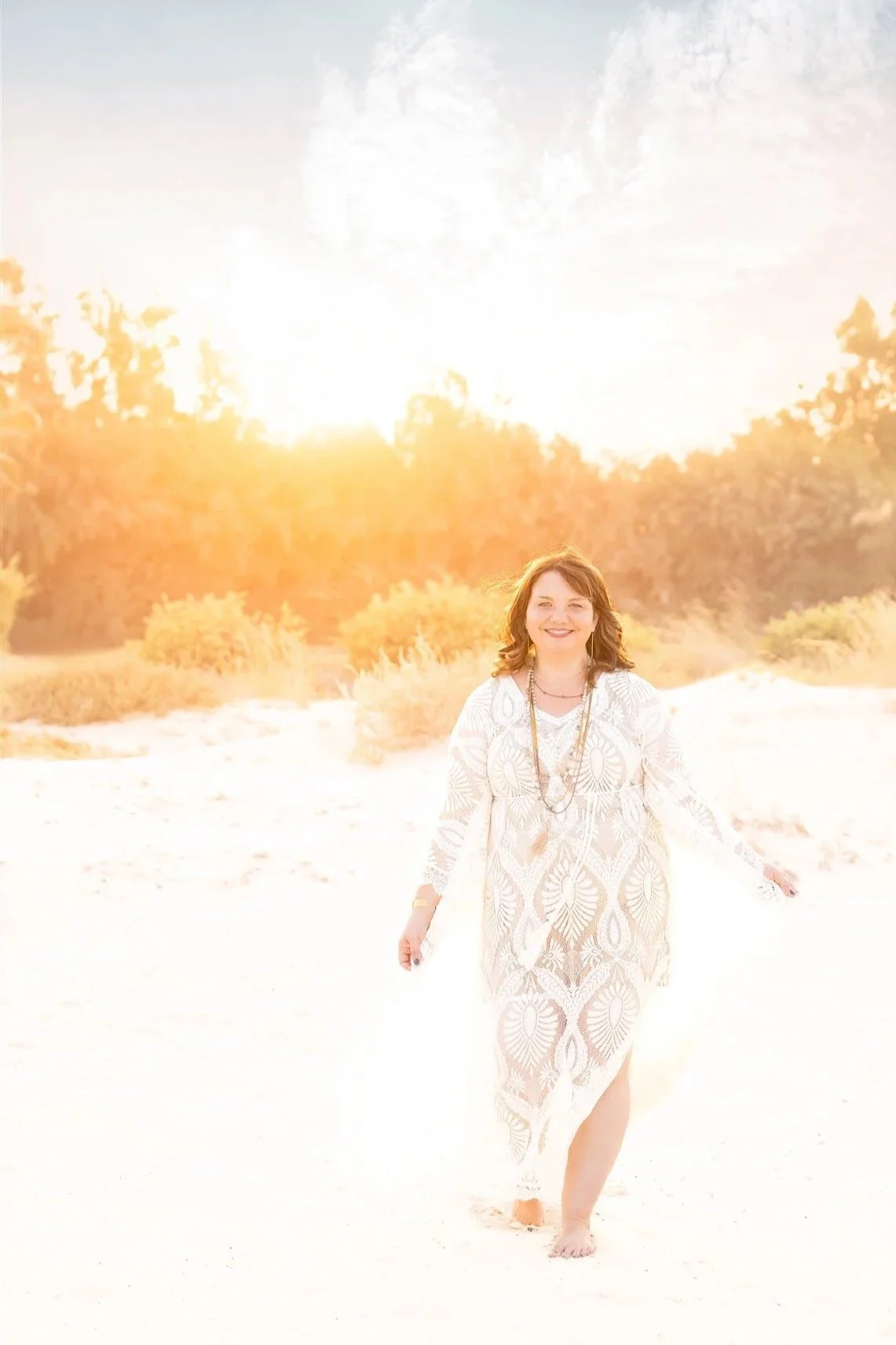 A woman smiling and walking barefoot on a sandy beach at sunset, wearing a white lace dress and necklaces, with trees and a bright sun in the background.
