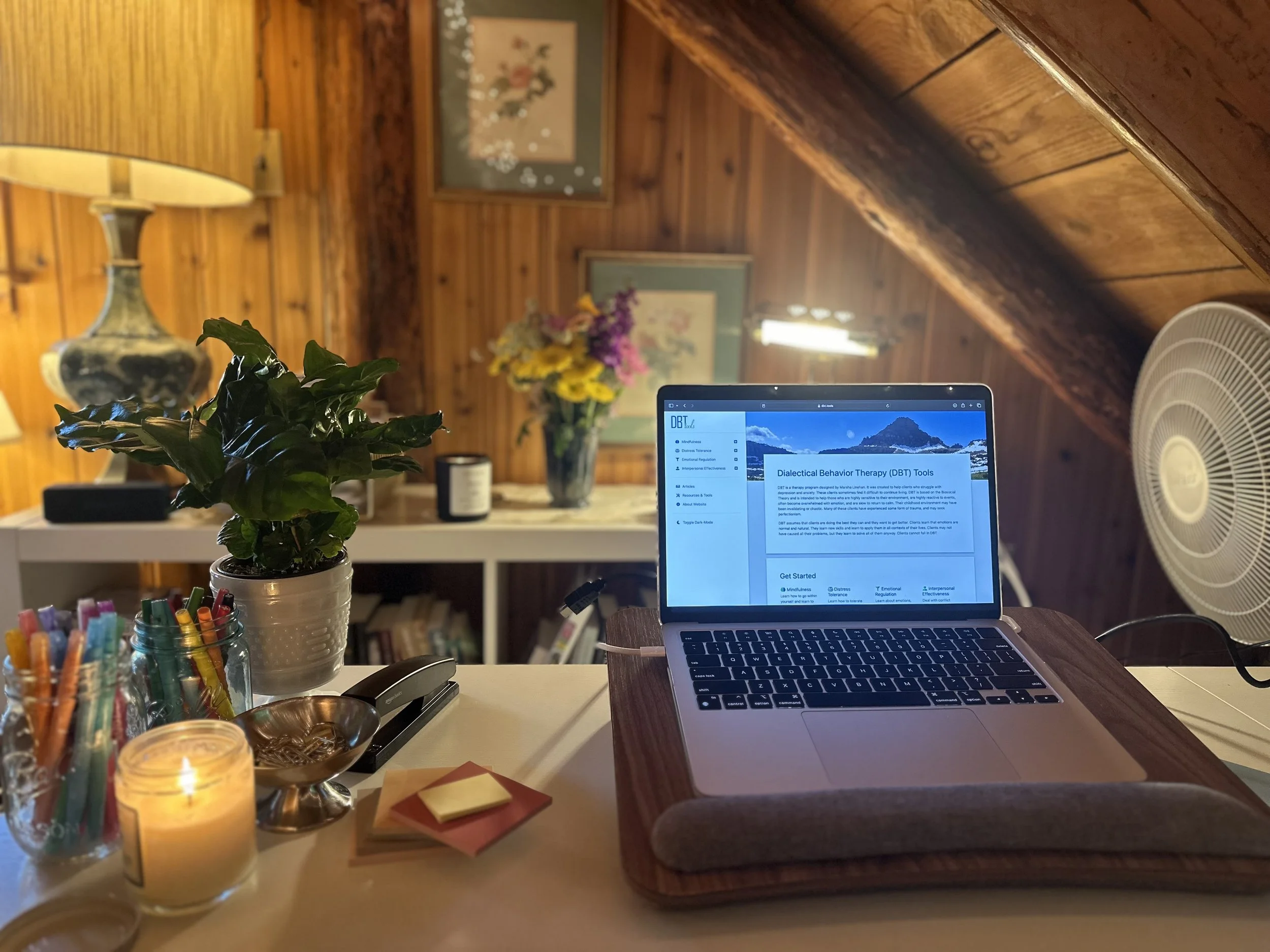 A cozy wooden-room home office with a laptop displaying Dialectical Behavior Therapy tools on a wooden desk, decorated with a large potted plant, colorful pens in a glass container, candles, and sticky notes. In the background, a Buddha tapestry, floral paintings, and a bouquet of flowers are visible.