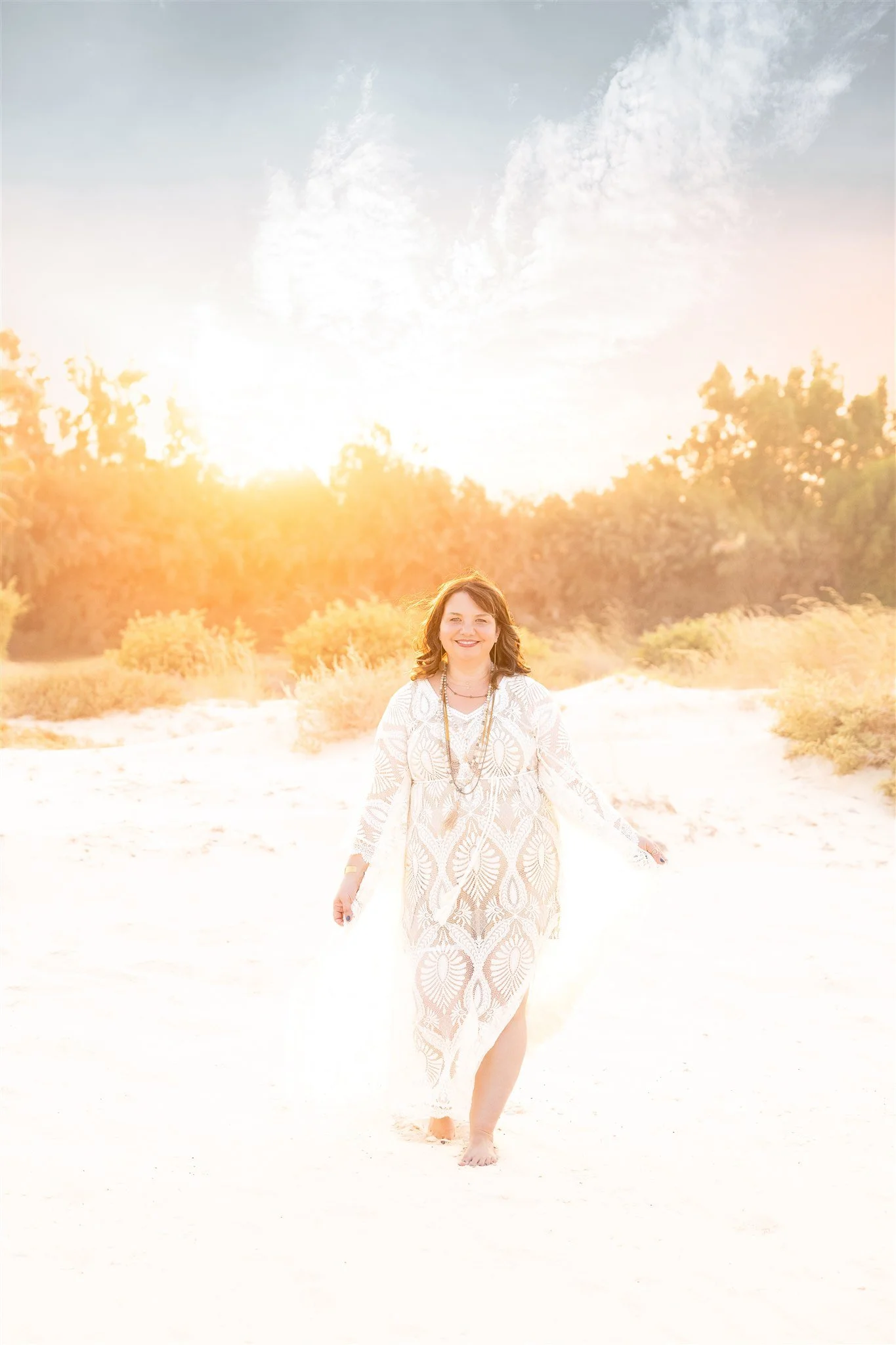A woman walking barefoot on a sandy beach at sunset, wearing a patterned white dress and smiling.