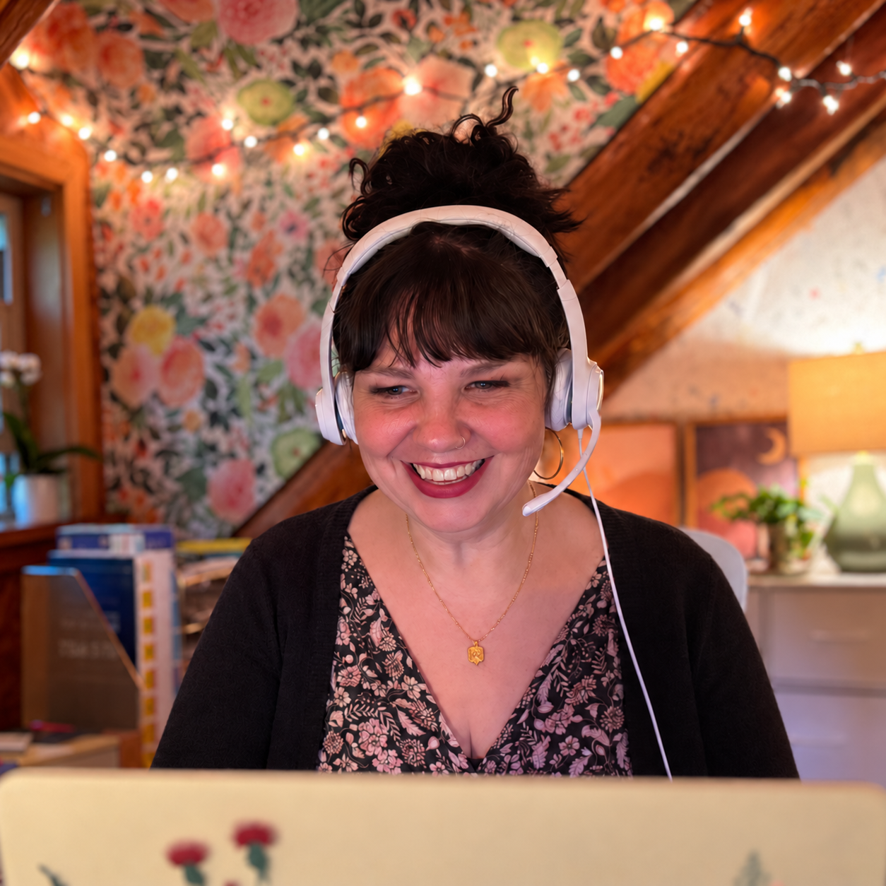 A woman therapist with short dark hair wearing a headset, smiling at a computer in a cozy room with floral wallpaper and warm lighting.