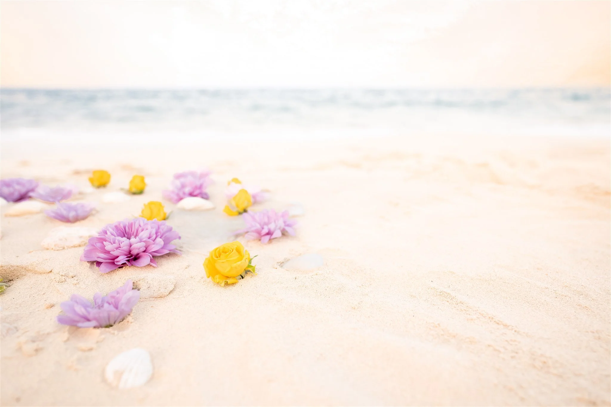 Colorful flowers, including pink, purple, and yellow, arranged on white sandy beach near the shoreline with ocean waves in the background.