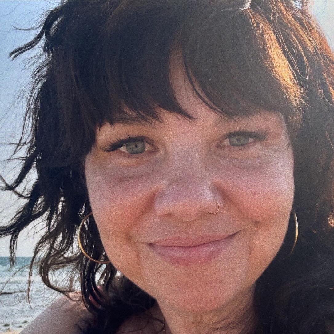 Close-up of a woman with dark curly hair, wearing hoop earrings and a nose ring, smiling on a beach with the ocean in the background during daylight.