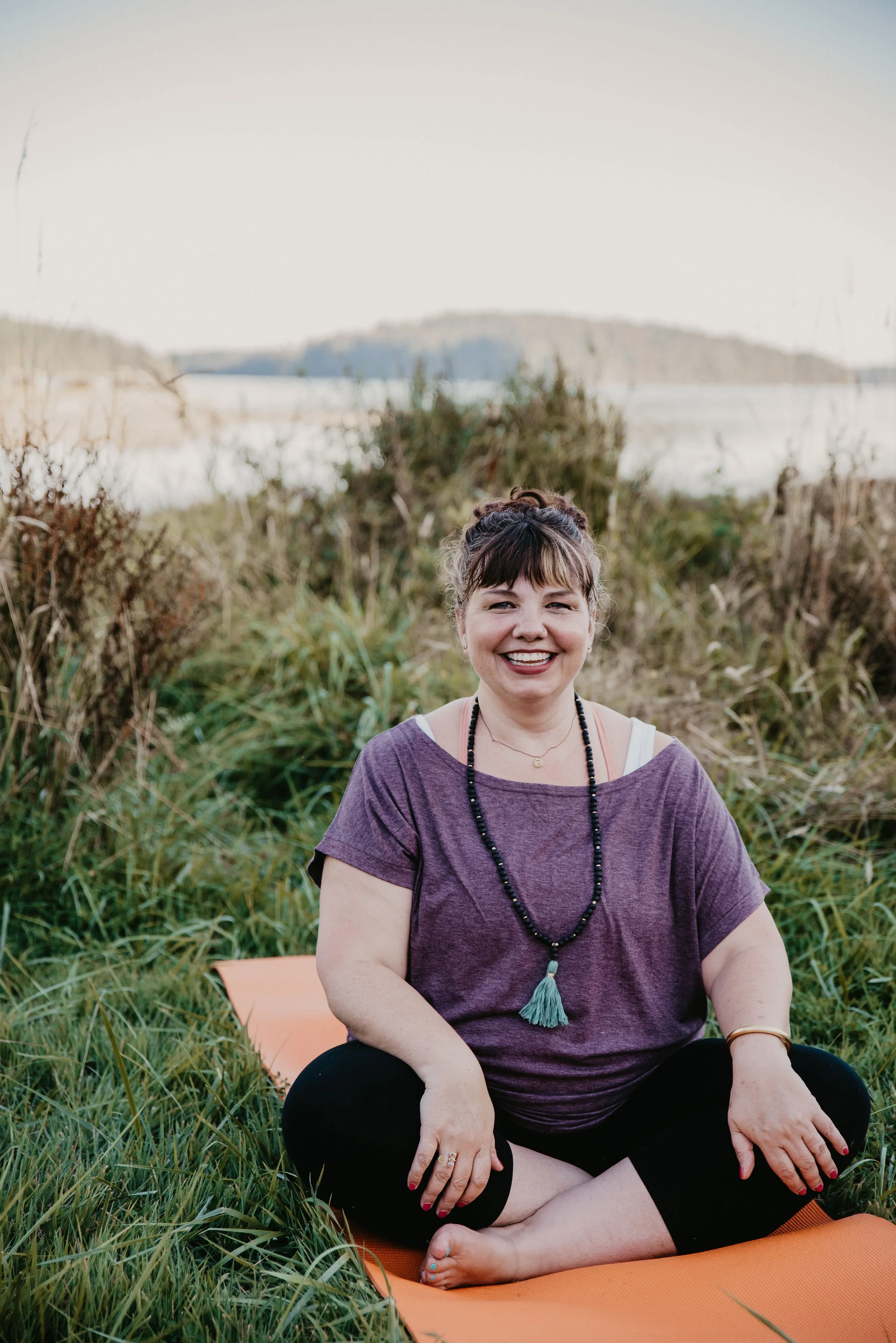 A woman with short hair and a big smile sitting cross-legged on an orange yoga mat outdoors in a grassy area near water, wearing a purple t-shirt, black pants, a beaded necklace, and a tassel necklace. There are plants and bushes in the background, and a blurred landscape with water and distant hills.