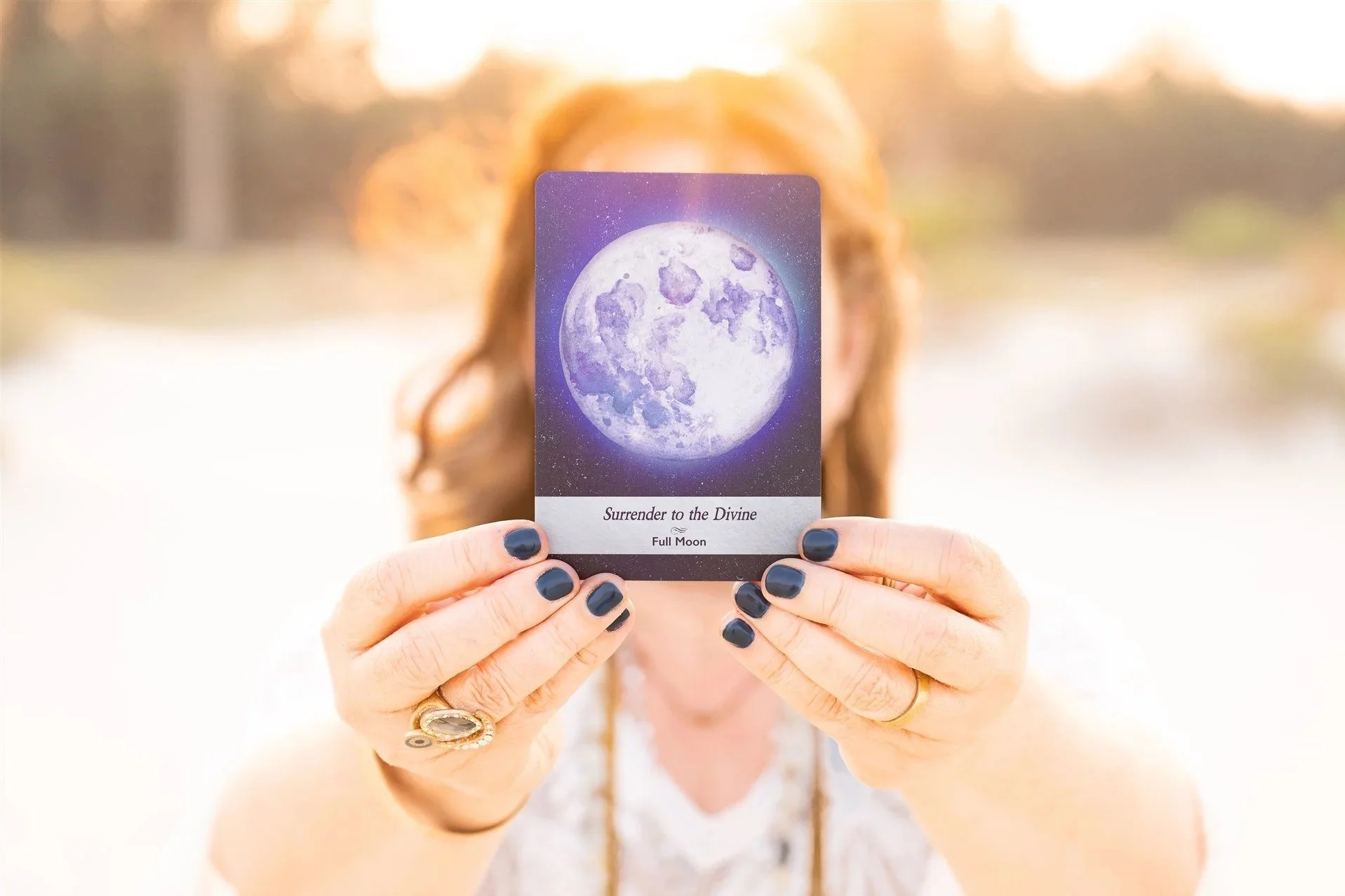 A person with dark nail polish and a ring holds up a card featuring a full moon illustration and the words "Surrender to the Divine" against a bright outdoor background.