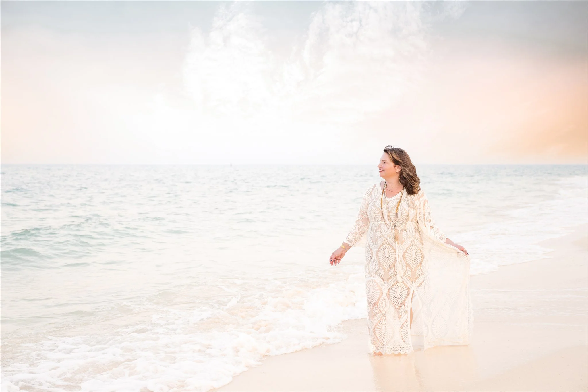 A woman in a long white dress with intricate patterns standing on a beach at sunset, facing right with a serene expression, holding her dress slightly with her right hand, beside the water with small waves.