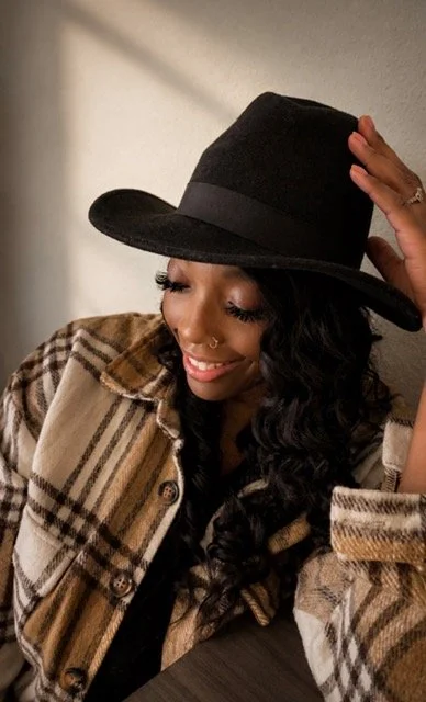 Deanna Goodwin, author, poet, and creator of The Pen She Carries, smiling in a black hat and plaid jacket.