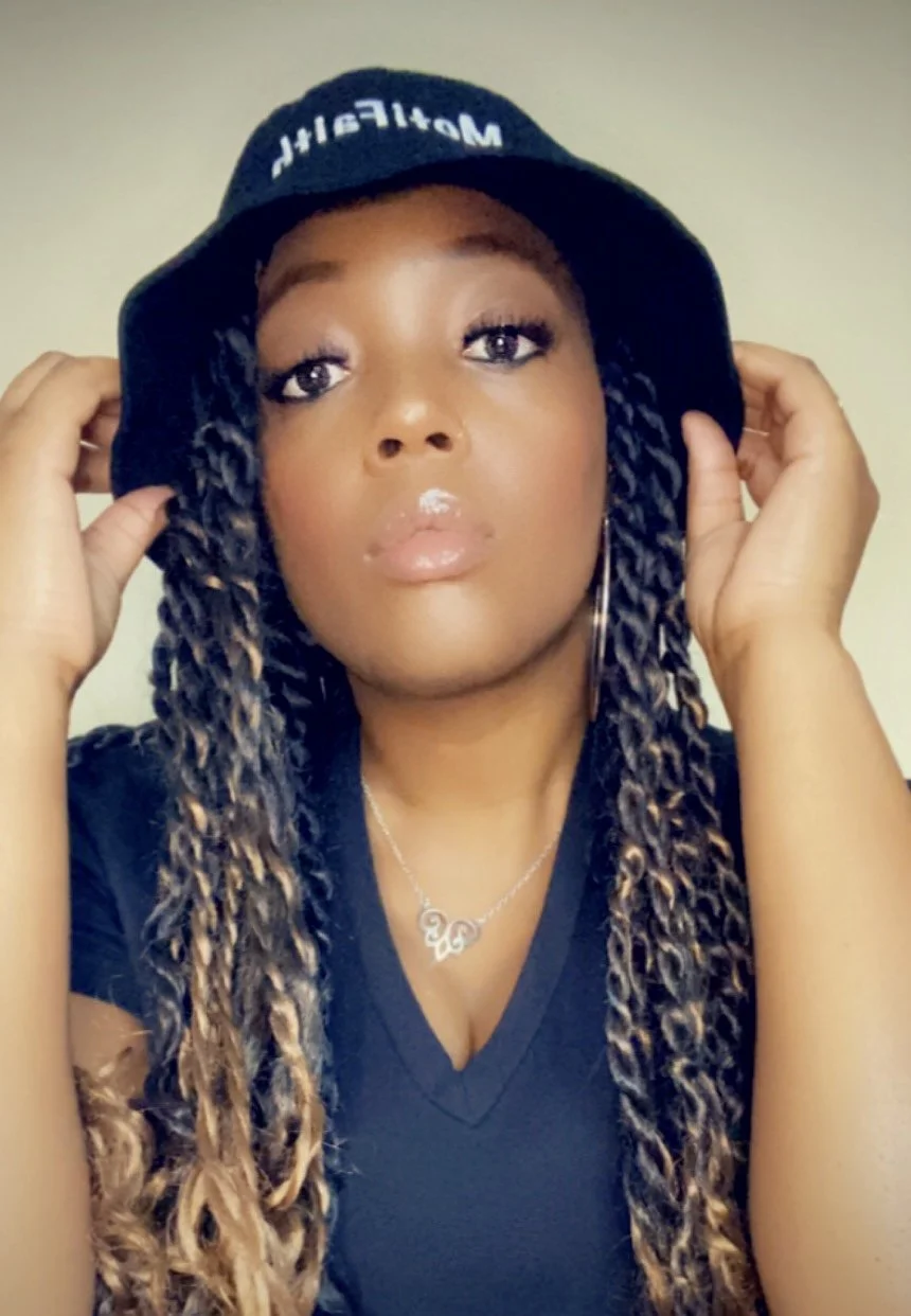 A woman wearing a black bucket hat with white lettering, black top, and gold twisted braids, holding the sides of her hat with both hands, looking into the camera.