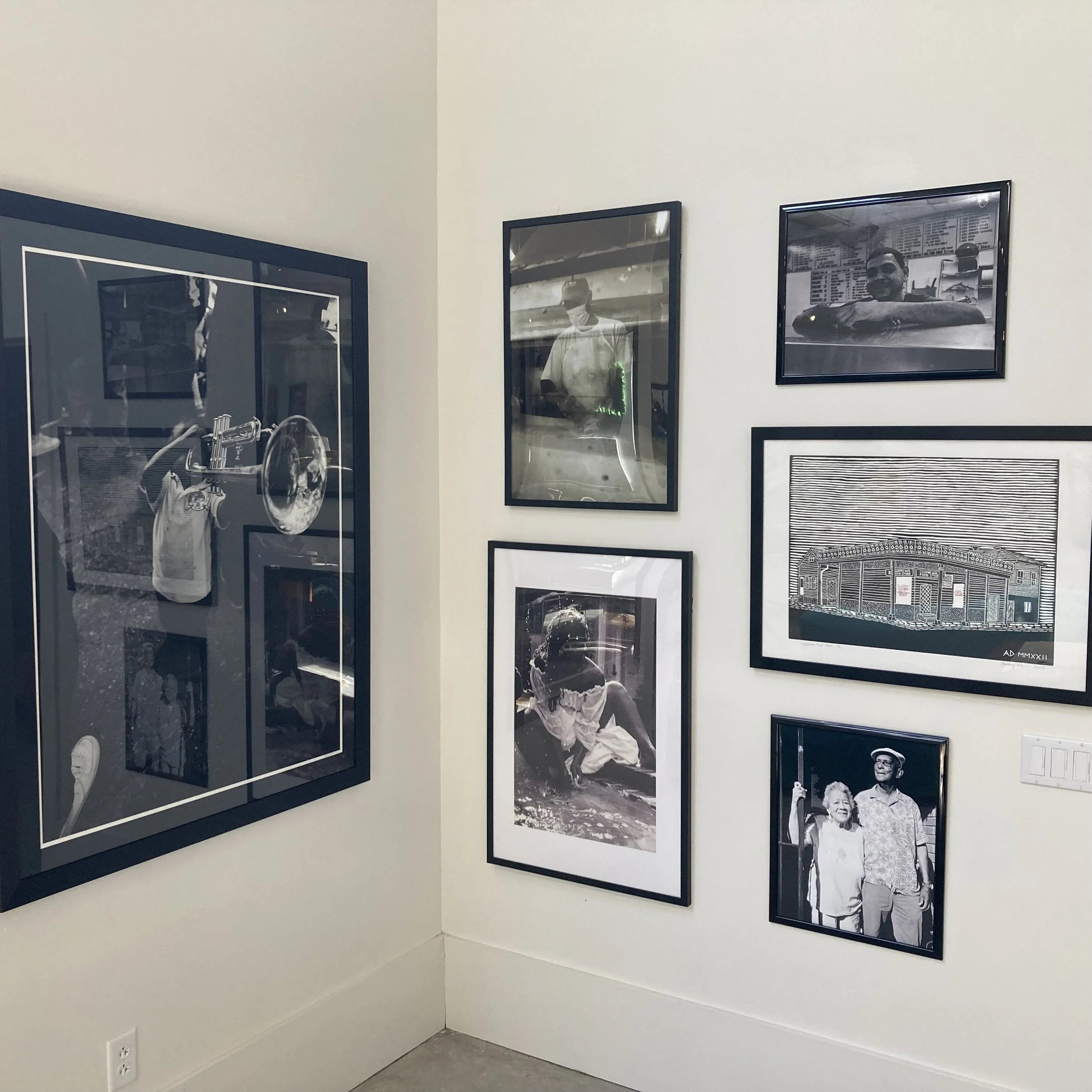Photo of black and white framed photographs on a white gallery wall, including images of a man with a hat and a smiling elderly couple, possibly at an exhibition or gallery