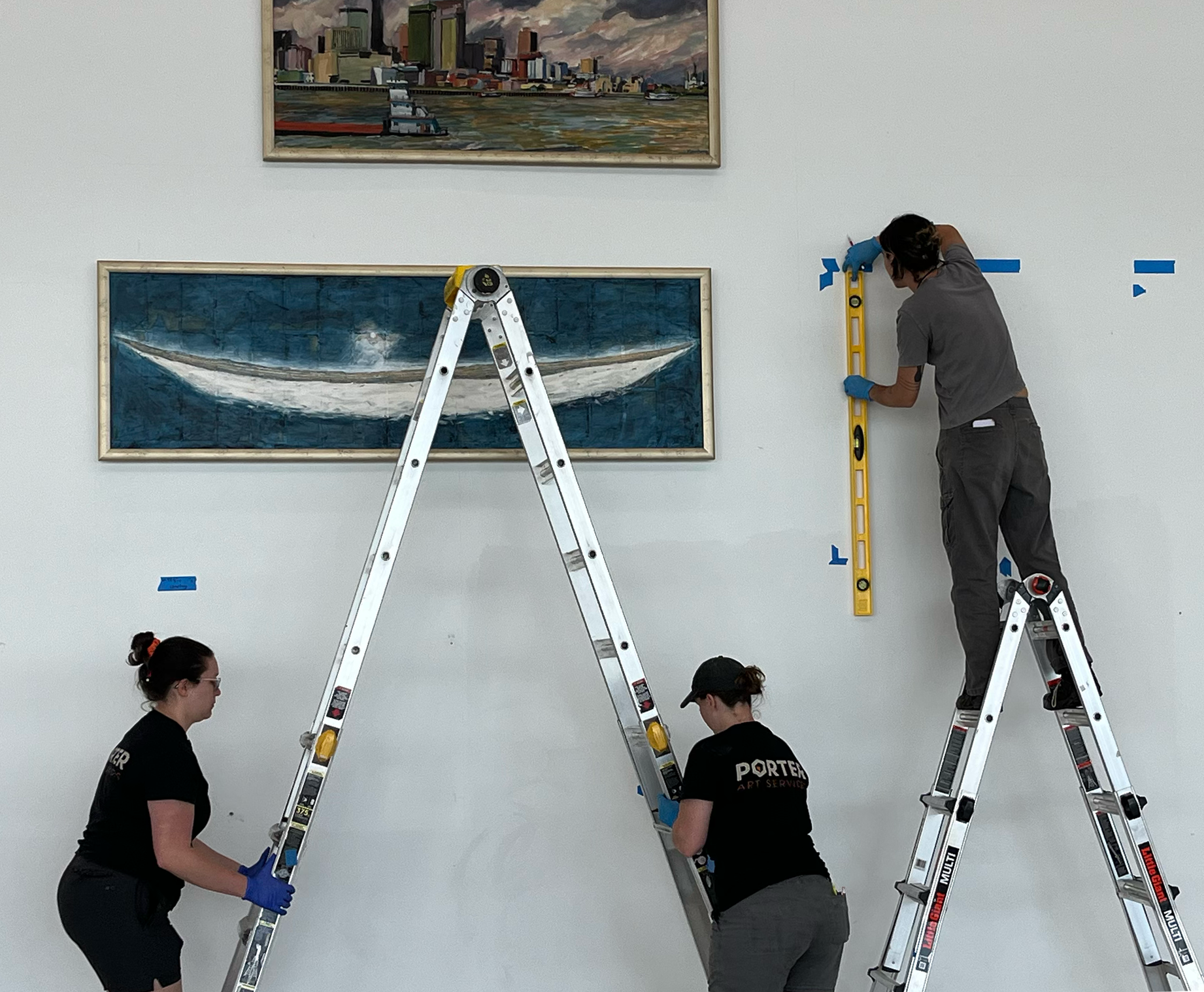 Museum staff members installing or preparing artwork on a white gallery wall. There are three women wearing black shirts and gloves, using ladders and tools. Two framed paintings are already hung on the wall, including a cityscape and a boat.
