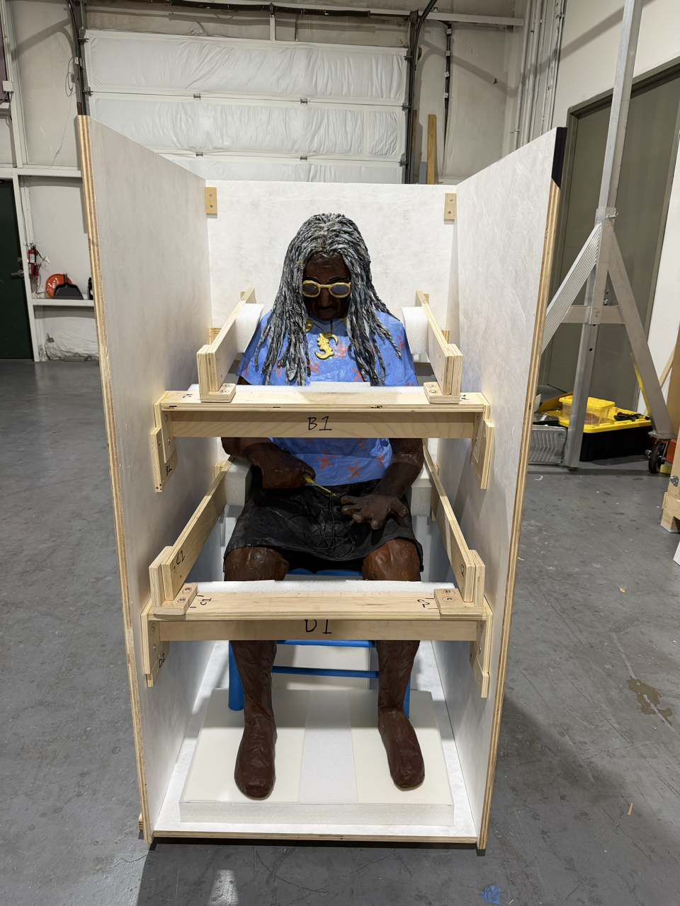 A person with dreadlocks, wearing glasses and a blue shirt, sitting in a wooden enclosure under construction with numbered beams and white walls.