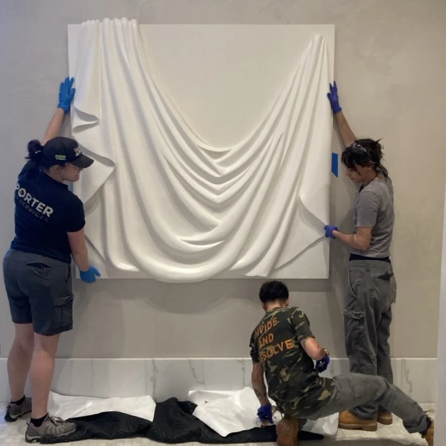 Three workers are installing a large white draped wall art piece, with two standing on either side pulling the fabric and one kneeling on the floor adjusting it, all wearing gloves.