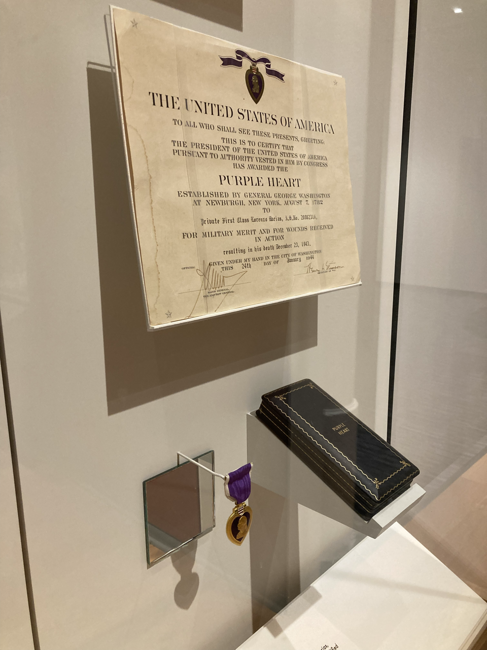 A framed certificate of honor for Private First Class Lorenzo Garias, awarded the Purple Heart, displayed on a wall. A medal with purple ribbon and a gold heart-shaped pendant hangs below, and a black case with gold edging rests on a white pedestal.