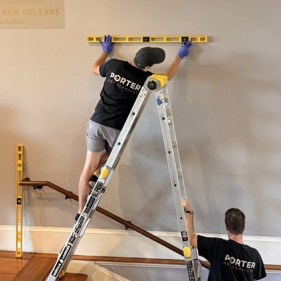 Two workers using a yellow level on a wall. One is standing on a ladder and the other is holding the ladder steady, both wearing black shirts with a company logo, gloves, and a cap.