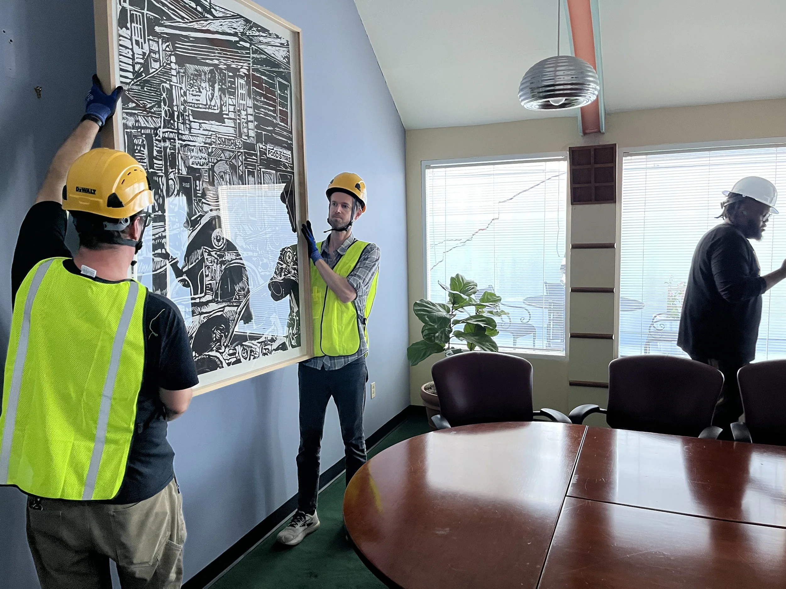 Three construction workers wearing yellow and white safety helmets and reflective vests are examining a large black-and-white artwork on the wall inside a room with a window and bookshelf.