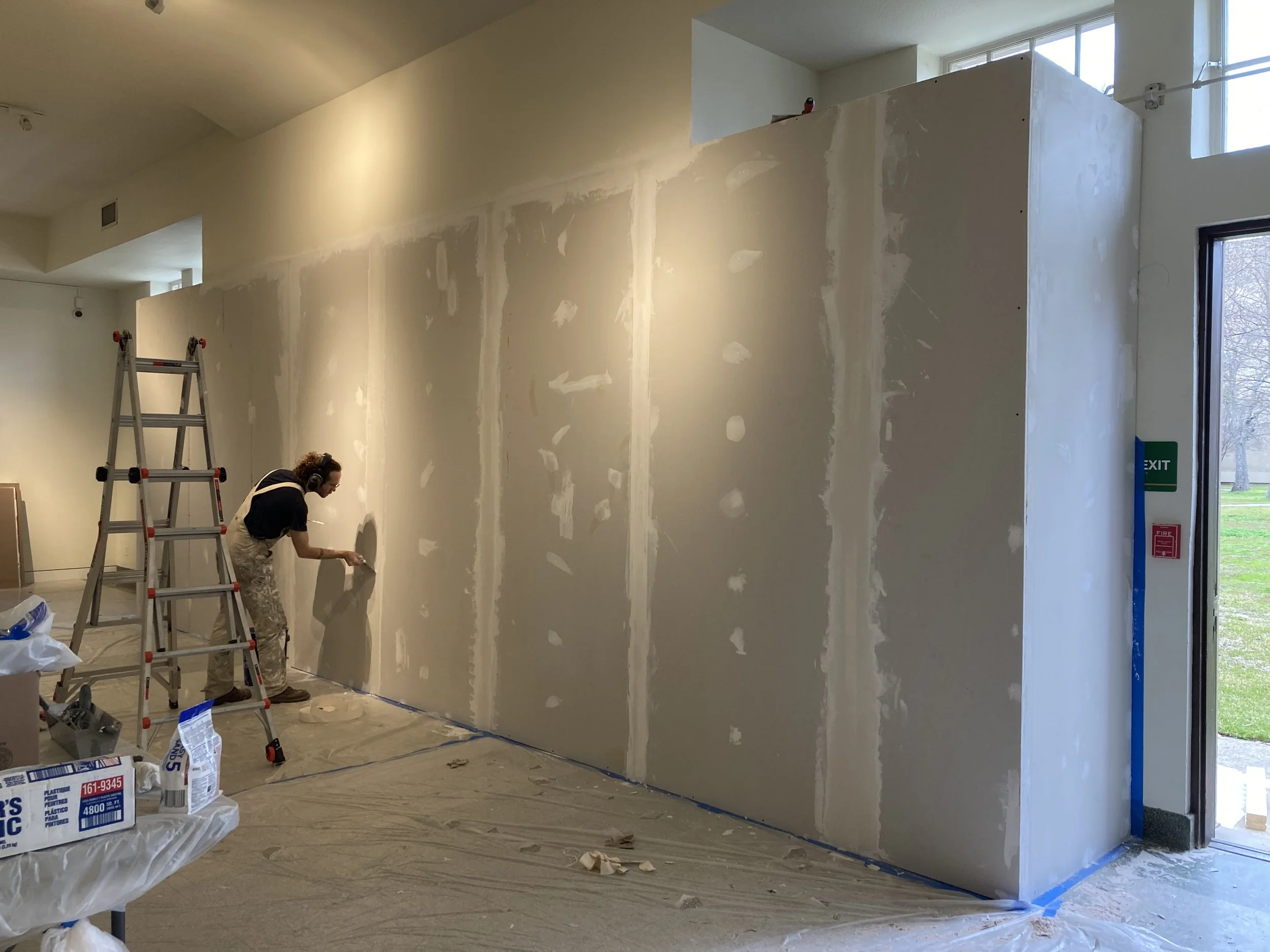 Interior of a building under renovation with drywall installation, a worker painting on a partially finished wall, a ladder, and construction materials on the floor.
