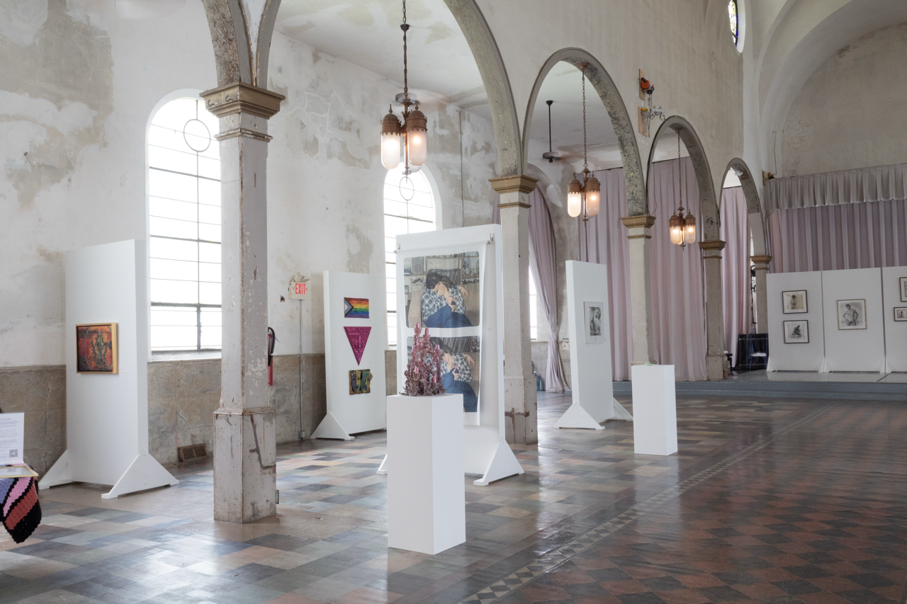 Interior view of an art gallery with white display panels and artwork, high ceilings with arches, large windows, pink curtains, and vintage hanging lights, in a spacious venue.