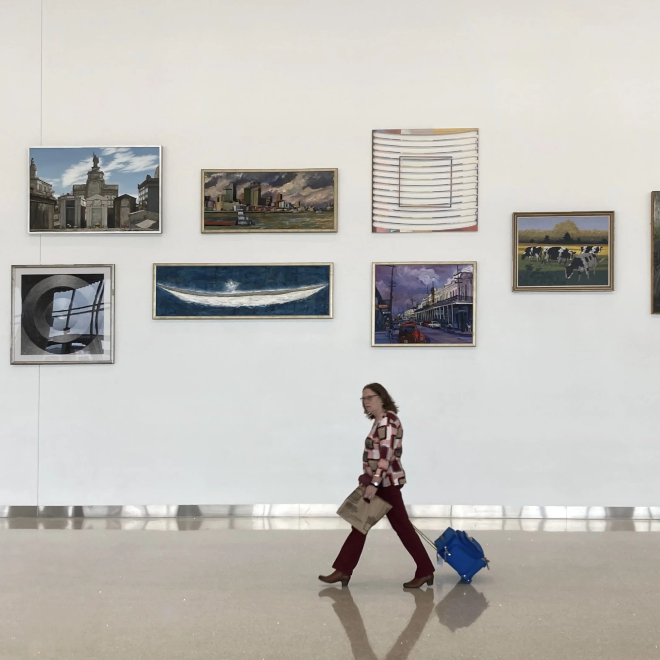 A woman walking through an art gallery carrying a shopping bag and pulling a rolling suitcase.