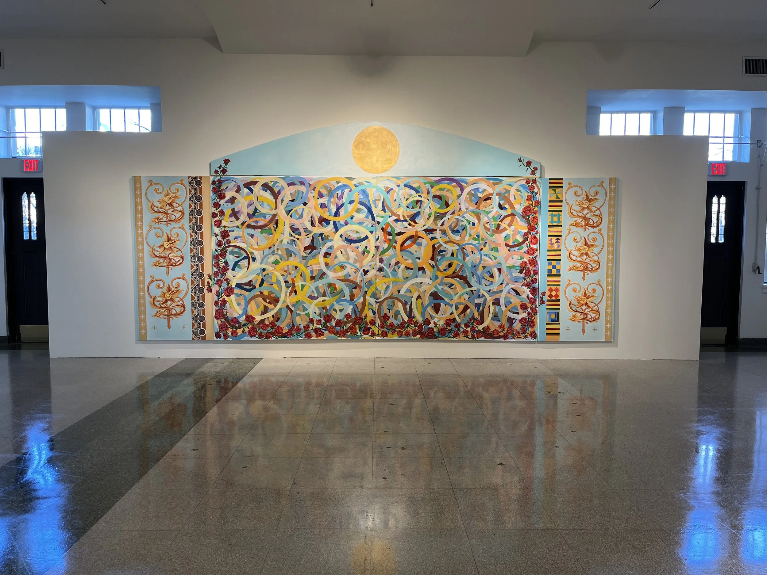 Large mural depicting a yellow sun above an abstract pattern of colorful intertwined circles, with decorative floral and geometric borders on a white wall in a building interior with windows and tiled floor.