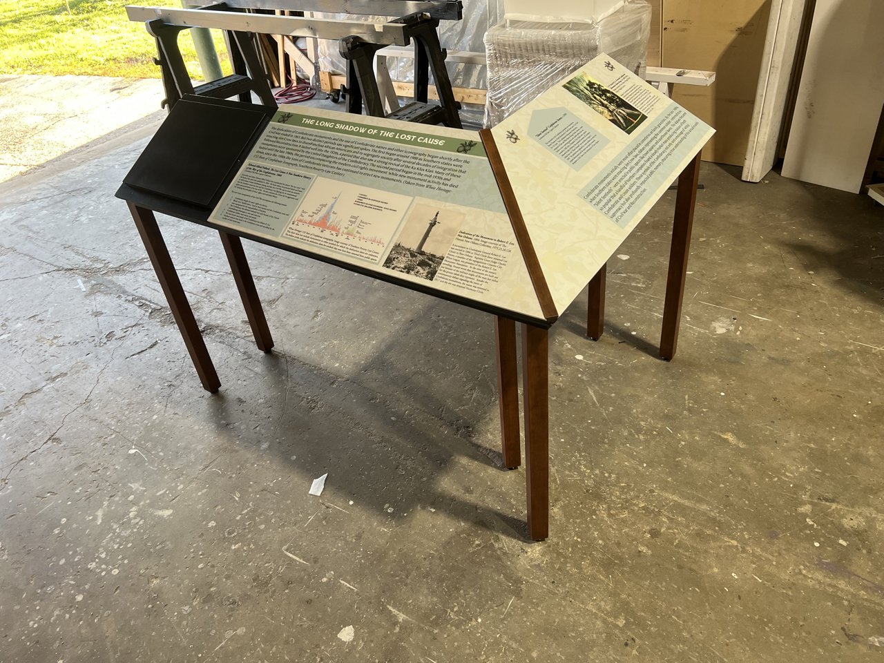 Information display panel with text and images about a historical event, set on a wooden stand in an indoor workshop or storage area with a concrete floor.
