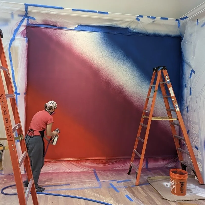 A person spray painting a gradient mural on a wall, with ladders and protective plastic covering the walls and floor.